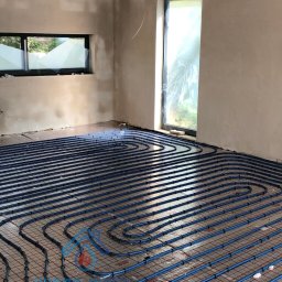 ADAM RADASZKIEWICZ Hydro-Instal - Niebieskie rury ogrzewania podłogowego ułożone na folii refleksyjnej z siatką w surowym wnętrzu, widoczne okna z folią ochronną.