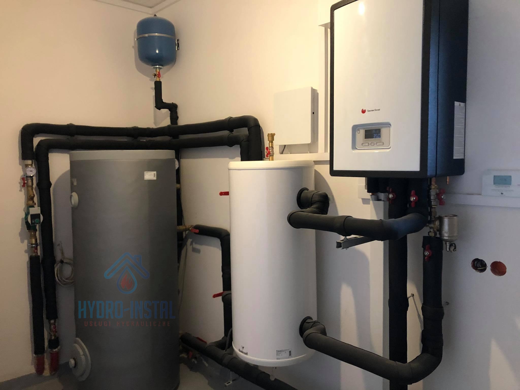 Nowoczesna kotłownia z białym kotłem gazowym Saunier Duval, szarym i białym zasobnikiem wody oraz niebieskim naczyniem przeponowym, rury w izolacji, widoczne przyłącza i zawory.