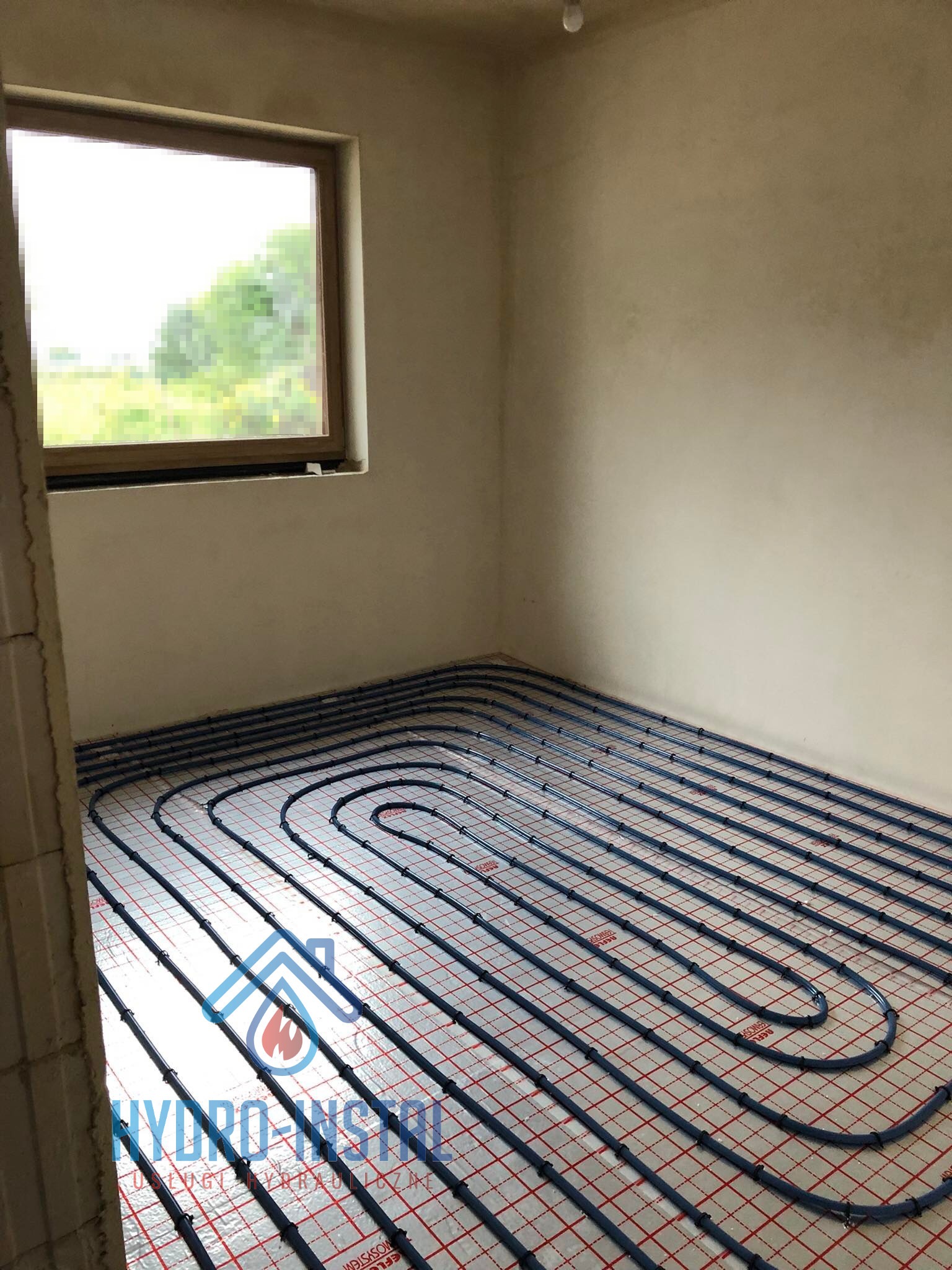 Niebieskie rury instalacji grzewczej ułożone równolegle na czerwonej siatce w surowym pomieszczeniu z oknem i widokiem na zieleń, w tle szare ściany.