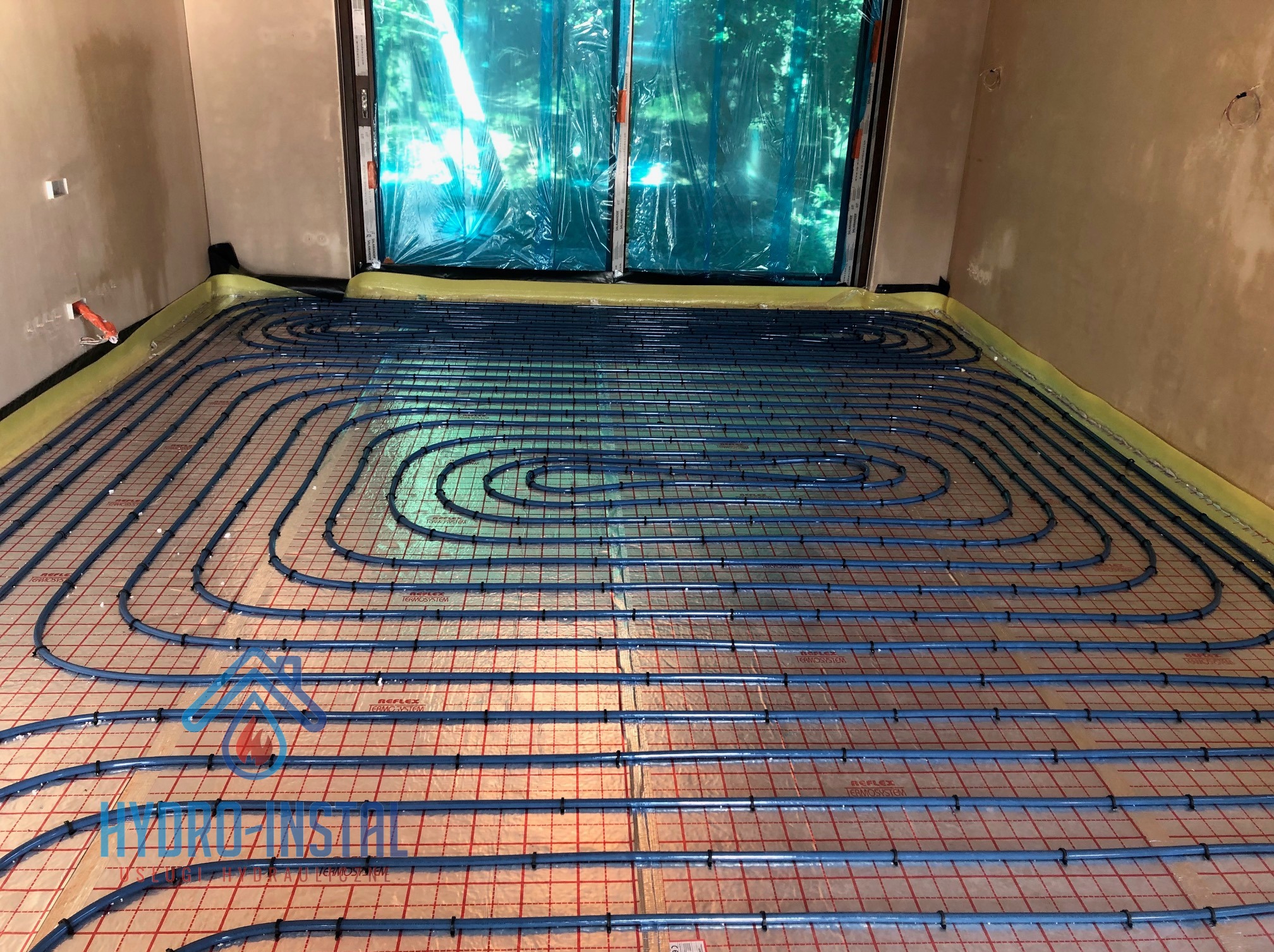 Niebieskie rury systemu ogrzewania podłogowego ułożone na folii z siatką, widoczne fragmenty ścian i okna zabezpieczonego folią malarską.