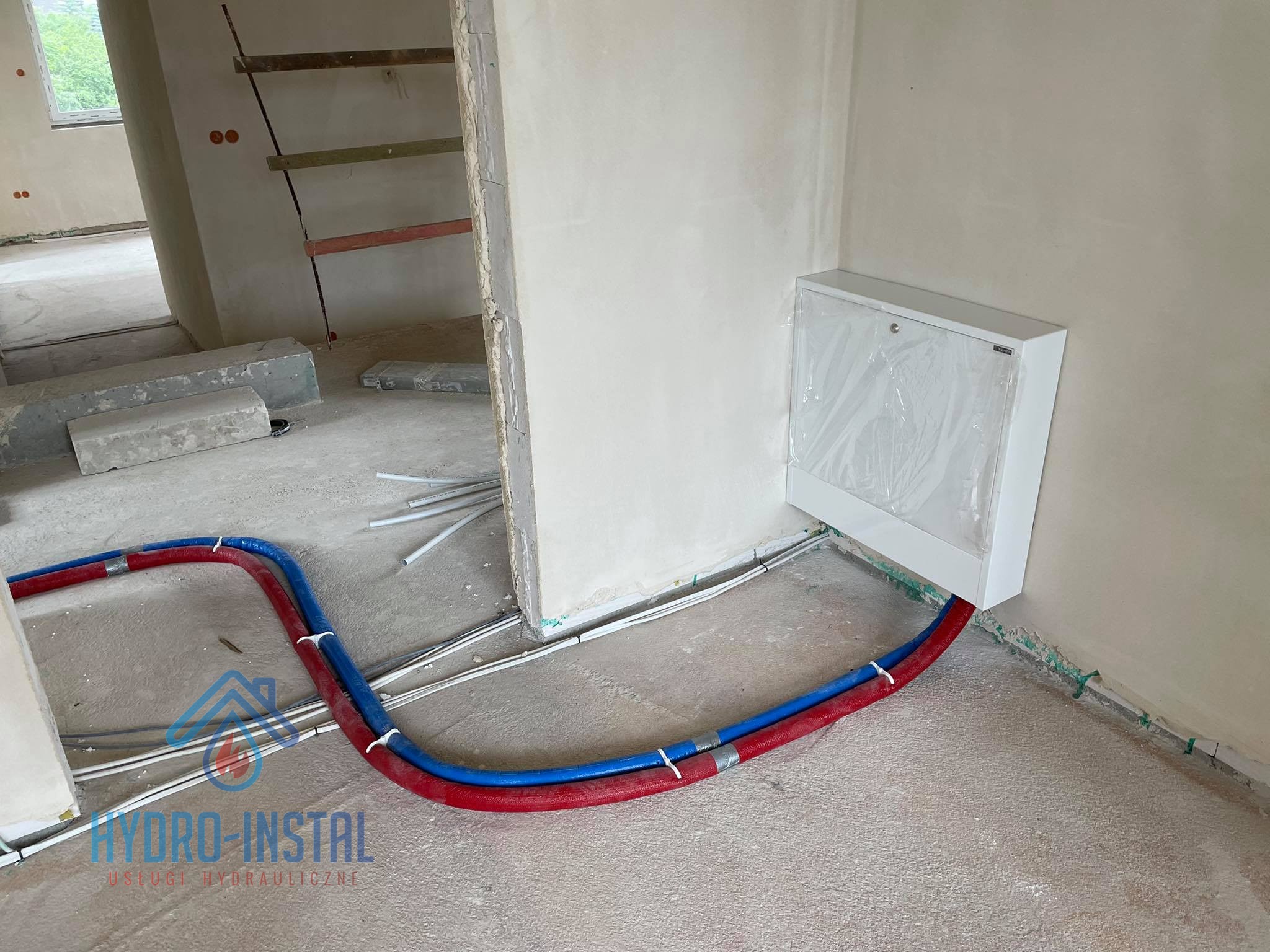 Instalacja ogrzewania podłogowego w trakcie realizacji we Wrocławiu, widoczne rury w izolacji czerwonej i niebieskiej, biała szafka rozdzielcza w folii ochronnej, surowy stan wykończenia wnętrza.