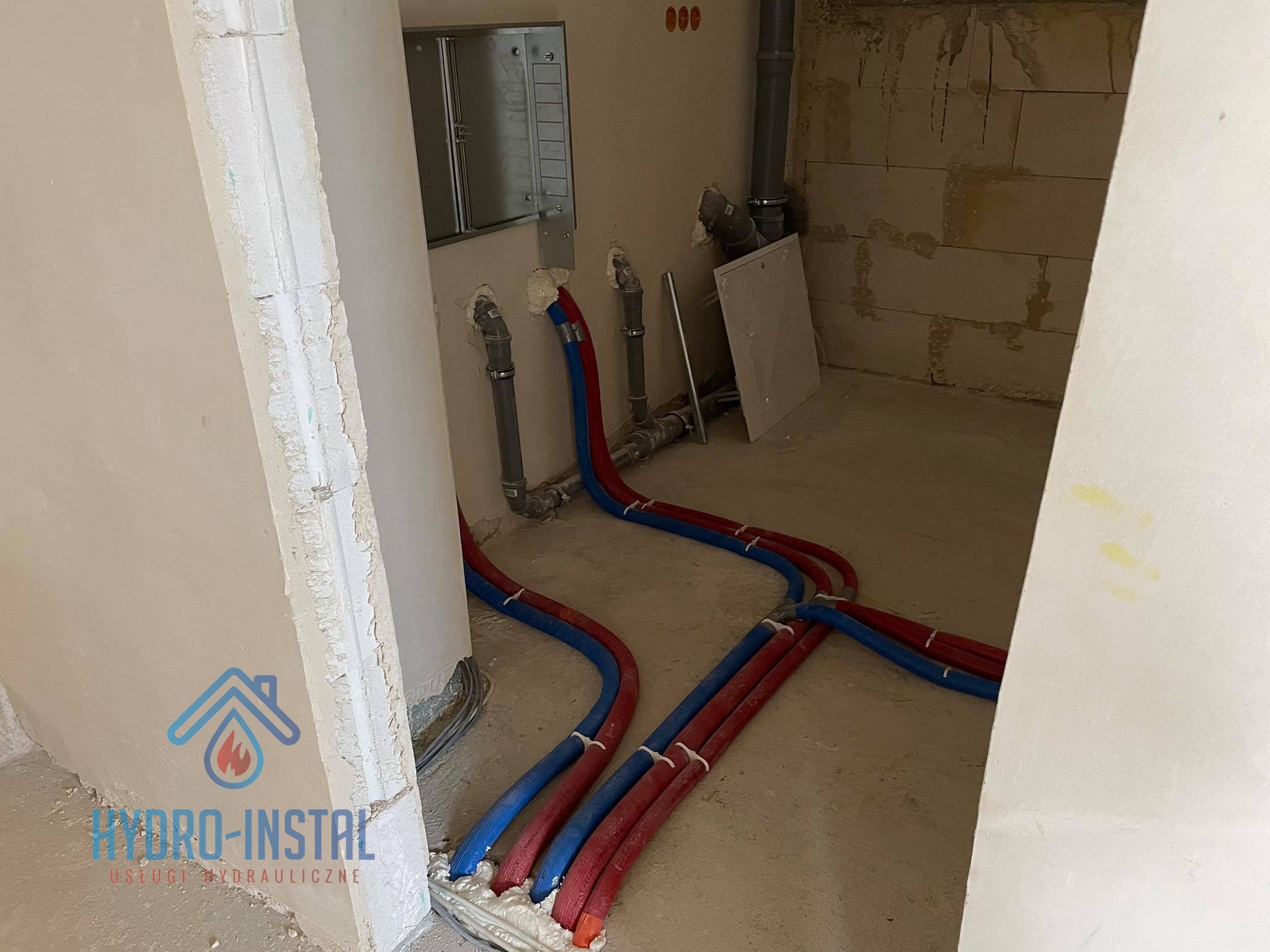 Wewnętrzne instalacje wodne z rur PEX w kolorze niebieskim i czerwonym, zabezpieczone pianką montażową, prowadzone w surowym stanie deweloperskim budynku, z widoczną skrzynką rozdzielczą na ścianie...