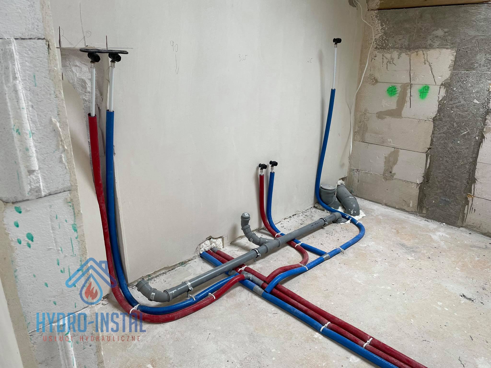 Surowe wnętrze w trakcie prac hydraulicznych: widoczne rury doprowadzające wodę, oznaczone kolorami czerwonym i niebieskim, oraz szare rury kanalizacyjne, zamontowane na ścianie i podłodze,...