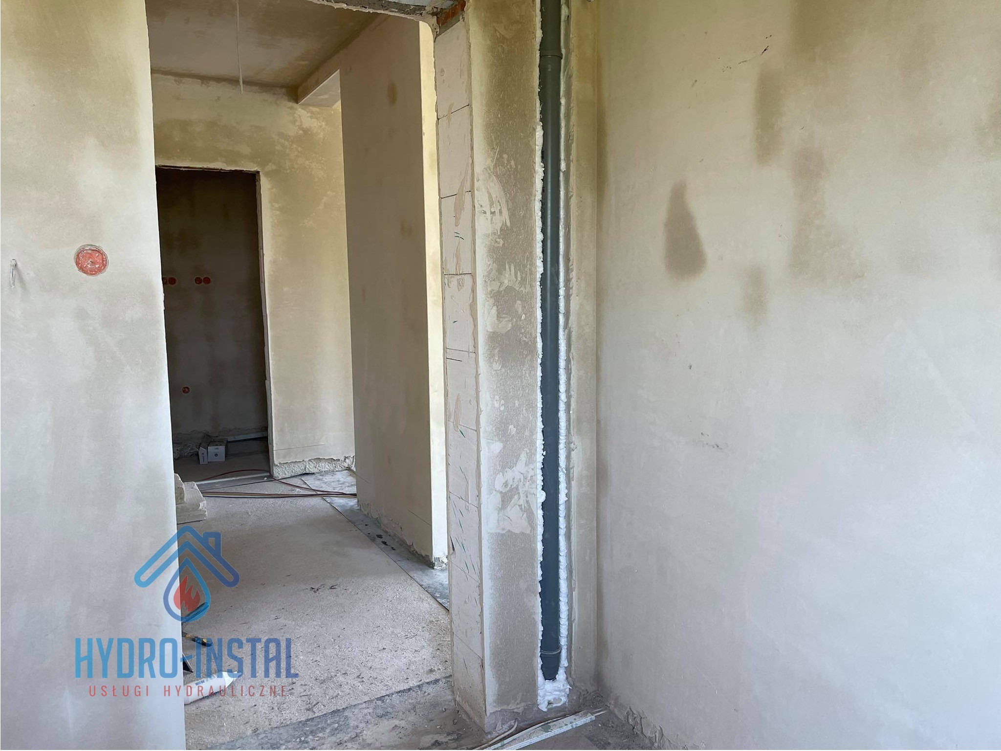 Szara rura kanalizacyjna pionowo zamontowana w wycięciu w ścianie, uszczelniona pianką montażową na tle surowego wnętrza w trakcie wykańczania.