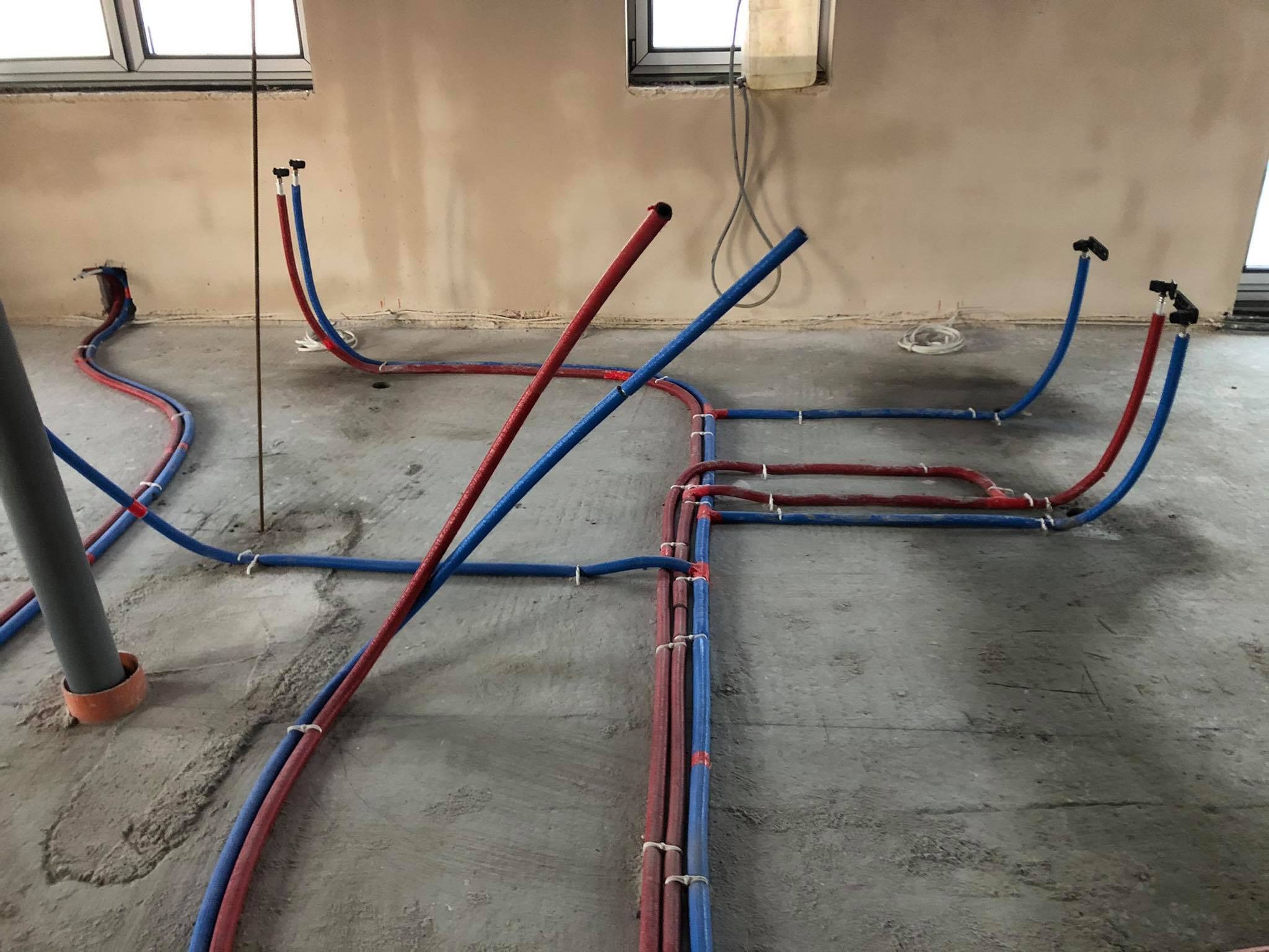 Instalacja ogrzewania podłogowego w trakcie montażu: rury PEX czerwone i niebieskie ułożone na surowym betonie, przymocowane plastikowymi uchwytami, widoczne otwory w podłodze na pionowe rury...