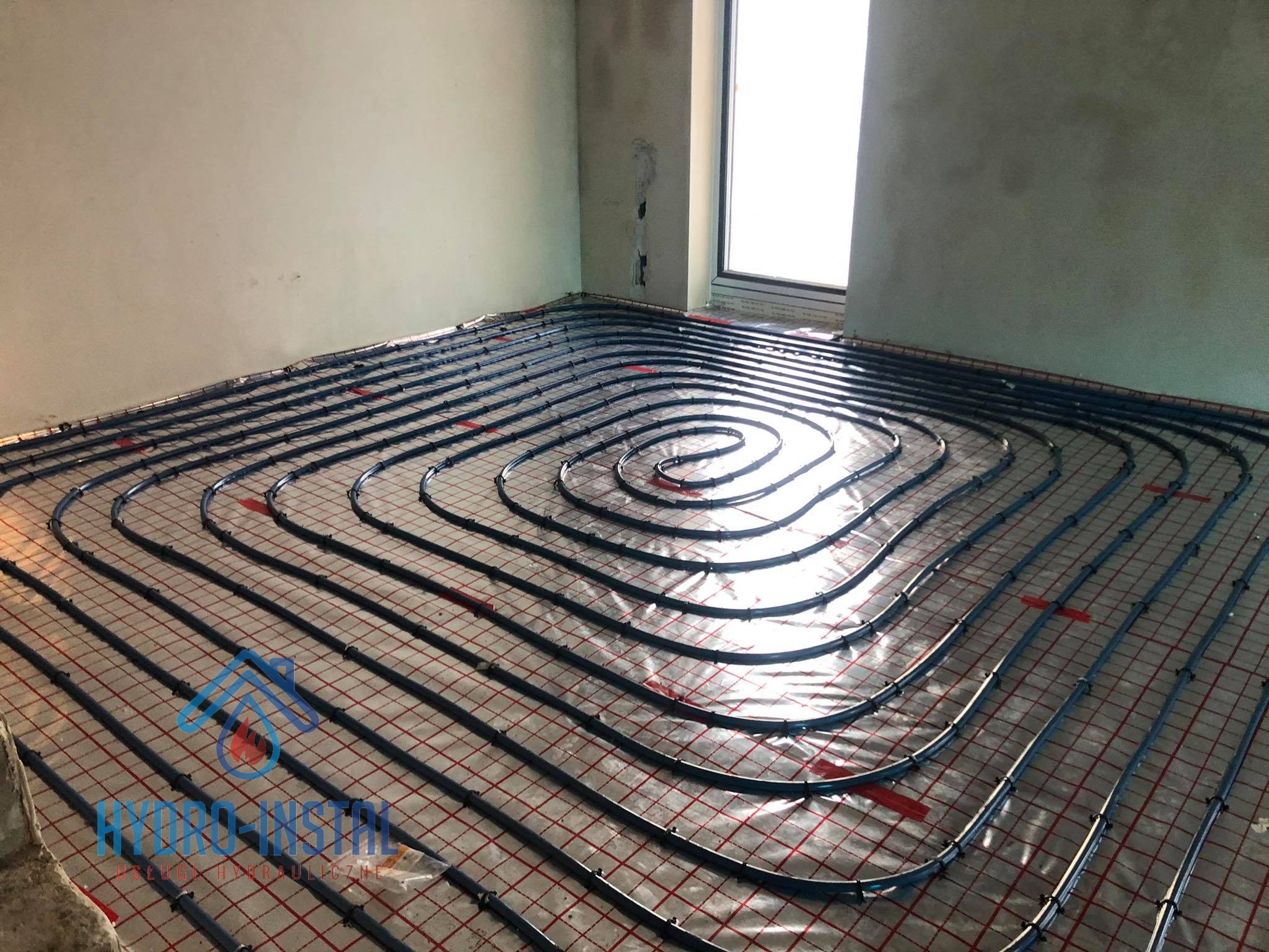 Niedokończona instalacja ogrzewania podłogowego z rurami ułożonymi na folii z siatką, widok z góry w surowym wnętrzu z oknem.
