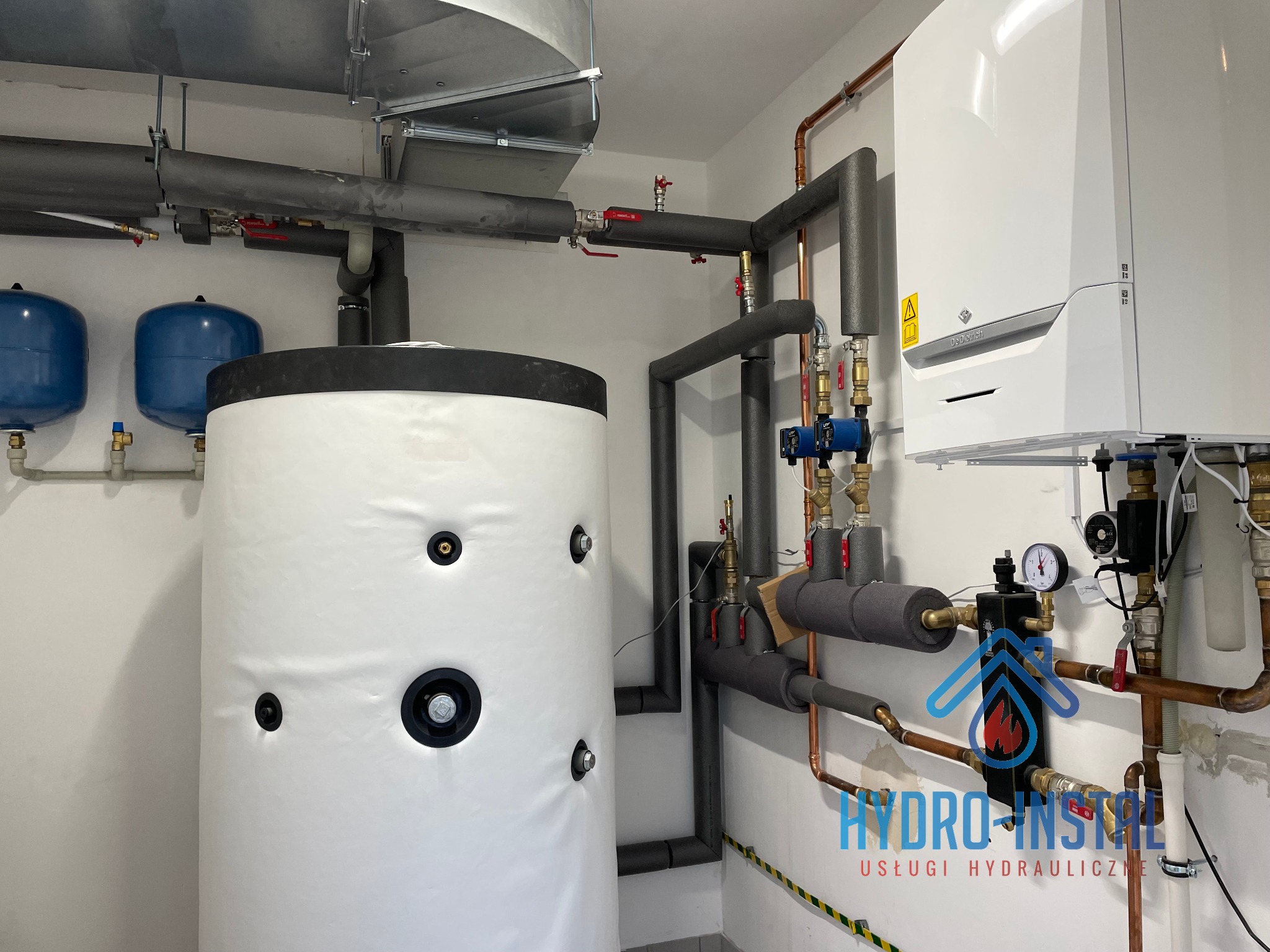 Biały zasobnik ciepłej wody użytkowej z izolacją, obok wiszący kocioł gazowy De Dietrich, rury miedziane i izolowane, dwa niebieskie zbiorniki wyrównawcze, widoczne logo firmy hydraulicznej...