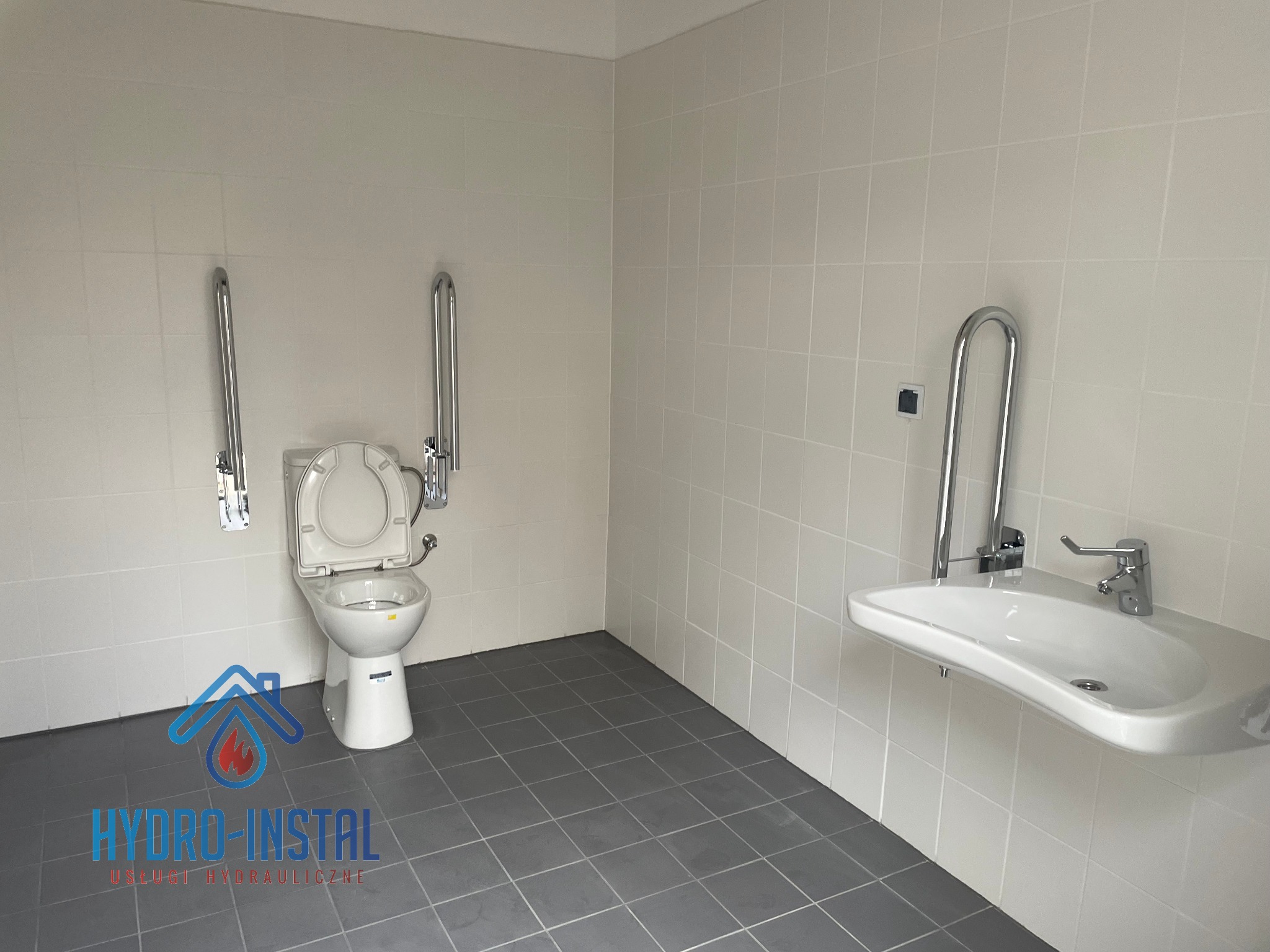 Wykończona łazienka dla niepełnosprawnych: białe płytki ścienne, szara podłoga, wisząca umywalka z chromowaną baterią i uchwytem, toaleta z uchwytami po obu stronach, logo firmy na podłodze.