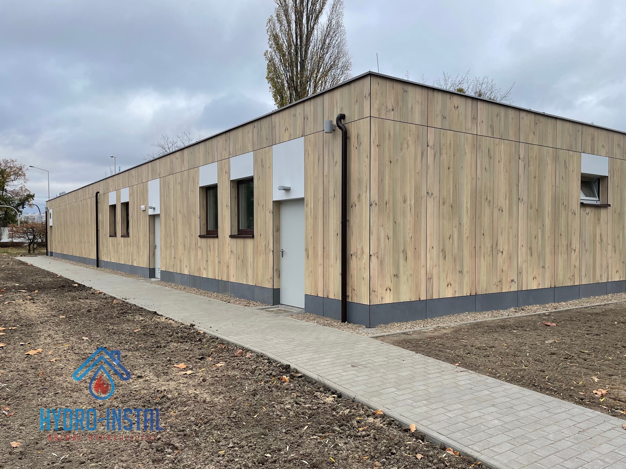 Zewnętrzny widok budynku socjalno-sportowego z drewnianą elewacją, szarą podmurówką i chodnikiem z kostki brukowej, wykonany w pochmurny dzień. Widoczne okna i drzwi wejściowe. Na pierwszym planie...