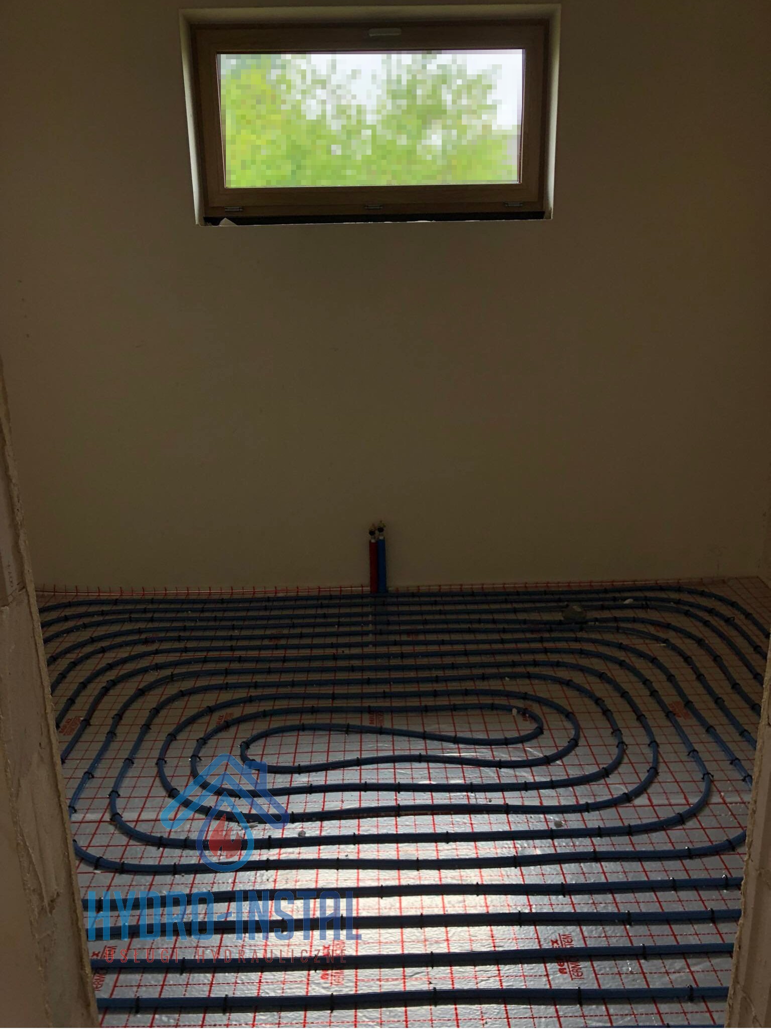 Nisko położone okno nad instalacją ogrzewania podłogowego z rurkami w kolorze niebieskim ułożonymi na srebrnej folii z czerwoną siatką.