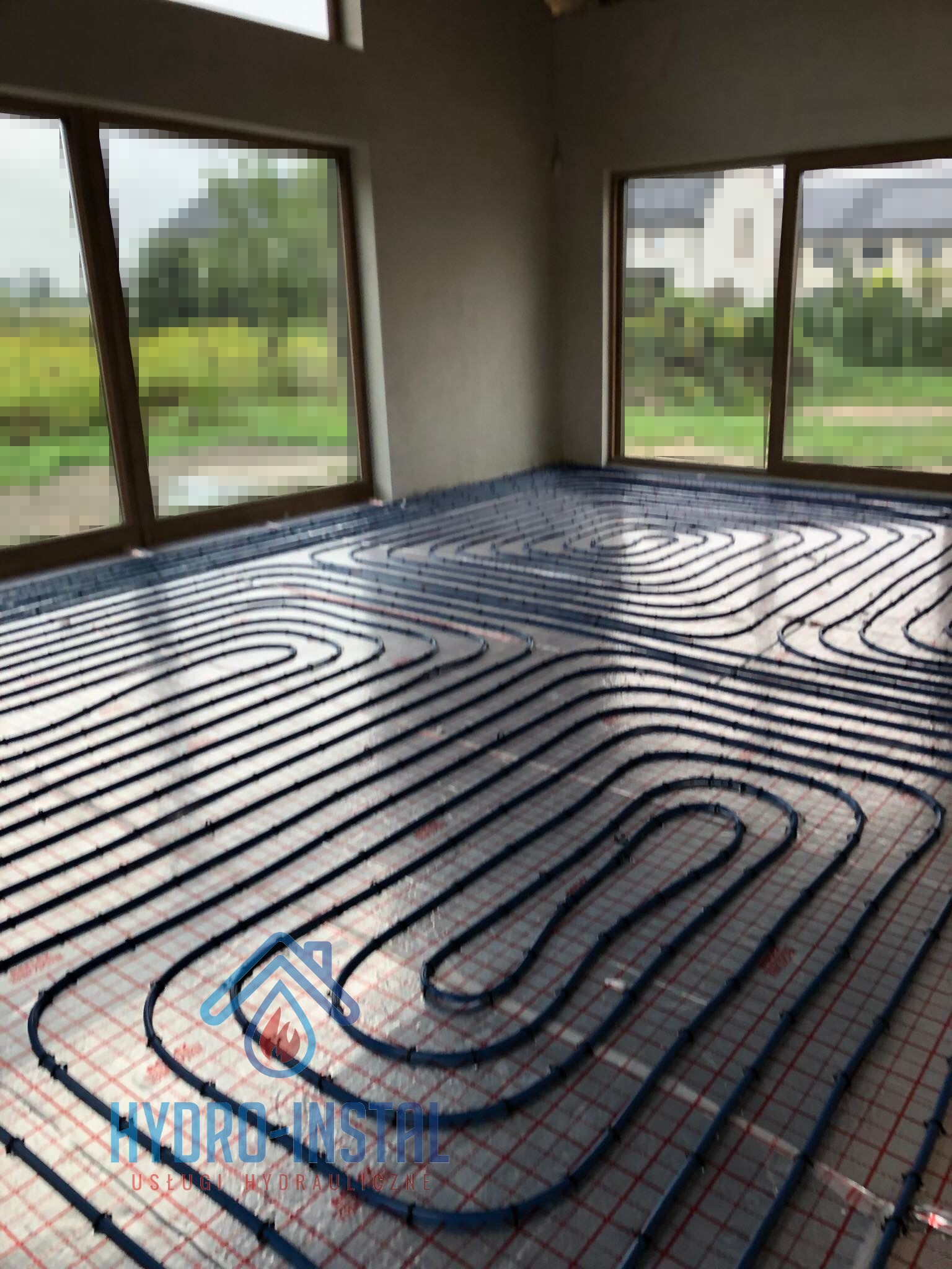 Rozłożone rury ogrzewania podłogowego na folii izolacyjnej z siatką, widok z góry na pomieszczenie z dwoma oknami i surowymi ścianami.