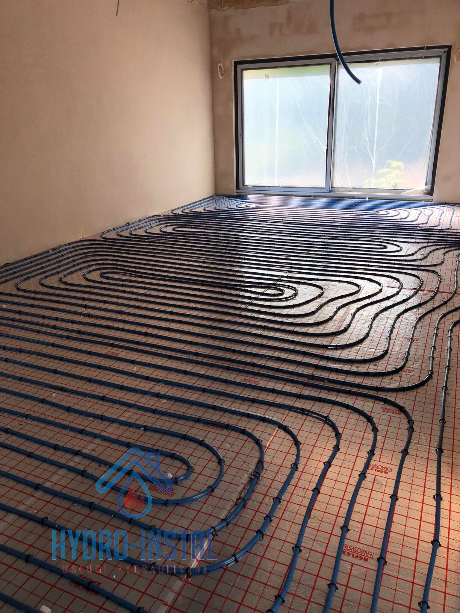 Niebieskie rury systemu ogrzewania podłogowego ułożone na folii z czerwoną siatką w surowym pomieszczeniu z oknem zabezpieczonym folią.