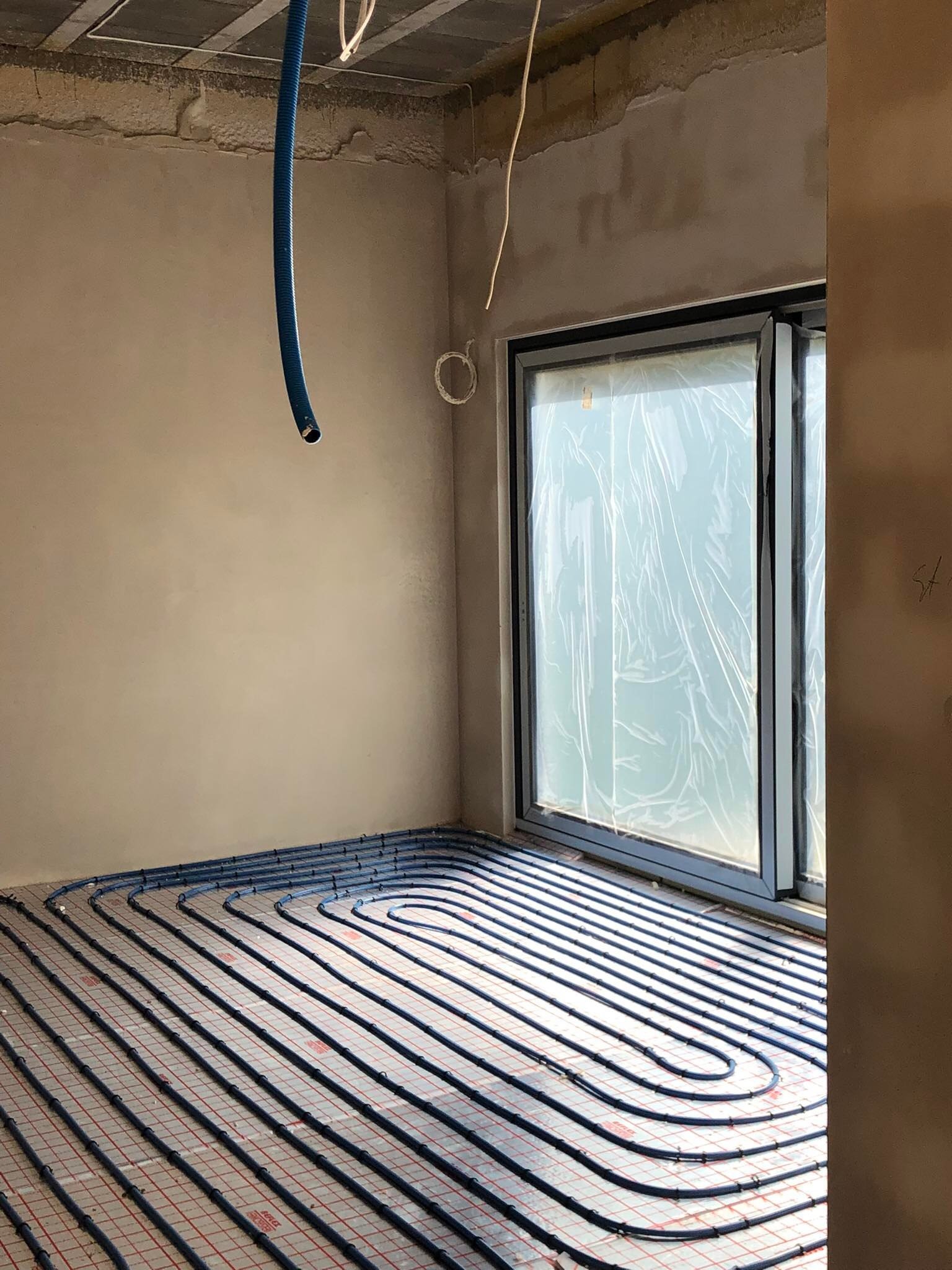 Niebieskie rury ogrzewania podłogowego ułożone na siatce w surowym pomieszczeniu z oknem zabezpieczonym folią, wisząca rura doprowadzająca.