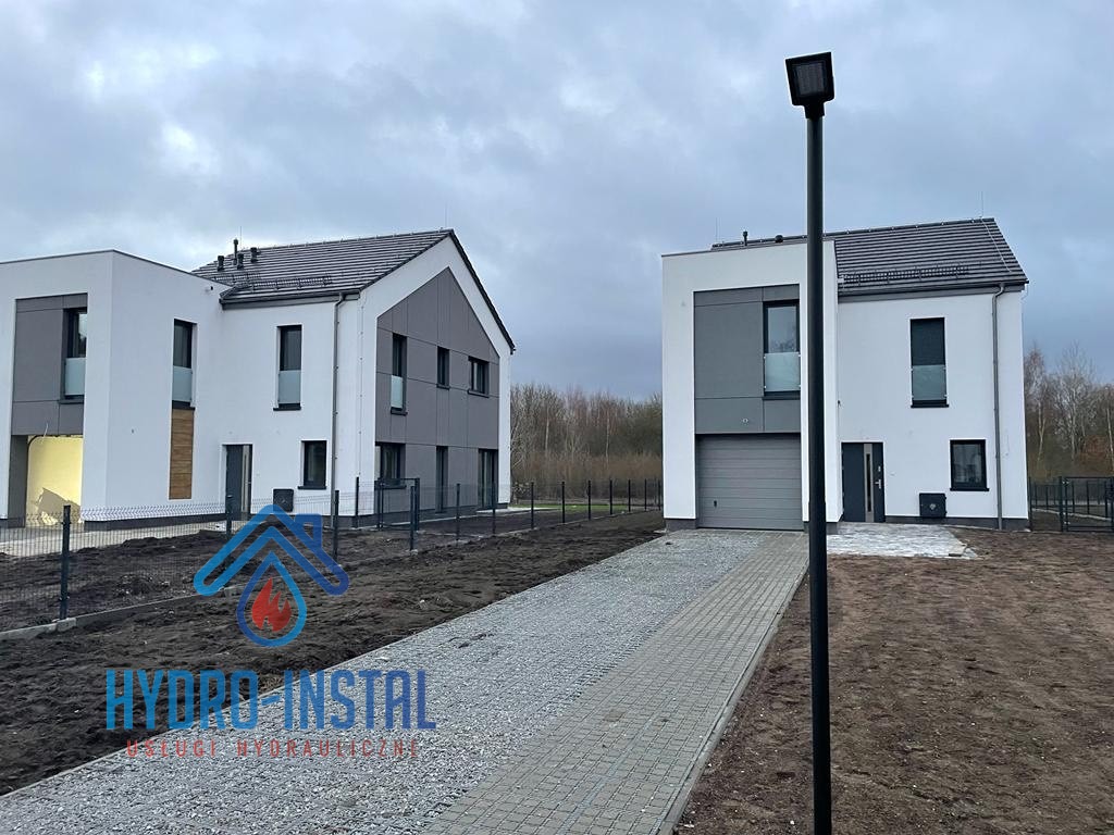 Dwa nowoczesne, dwukondygnacyjne domy jednorodzinne w zabudowie bliźniaczej, z widocznym logo firmy hydraulicznej na pierwszym planie, teren wokół domów jeszcze niezagospodarowany, szara kostka...
