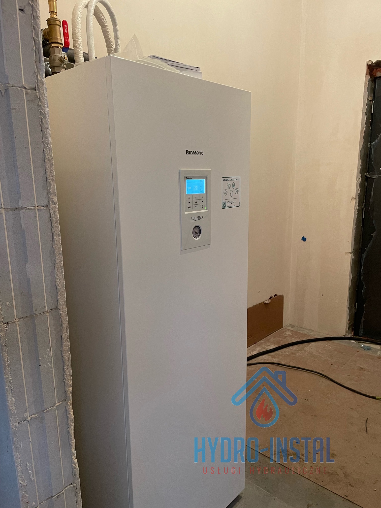 Biała pompa ciepła Panasonic Aquarea R32 z widocznym panelem sterowania, zamontowana w surowym pomieszczeniu z fragmentem ściany z betonu komórkowego i instalacjami hydraulicznymi.