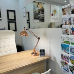 HARMONY FINANSE SPÓŁKA Z OGRANICZONĄ ODPOWIEDZIALNOŚCIĄ - Jasne biuro z drewnianym biurkiem, miedzianą lampką, białym krzesłem i ulotkami w tle, prawdopodobnie punkt obsługi klienta.