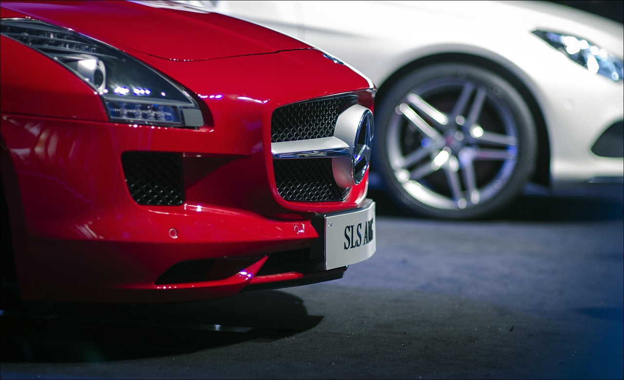 Czerwony sportowy samochód SLS AMG z widocznym grillem, logo Mercedes-Benz i reflektorami LED, obok biały samochód z aluminiowymi felgami.