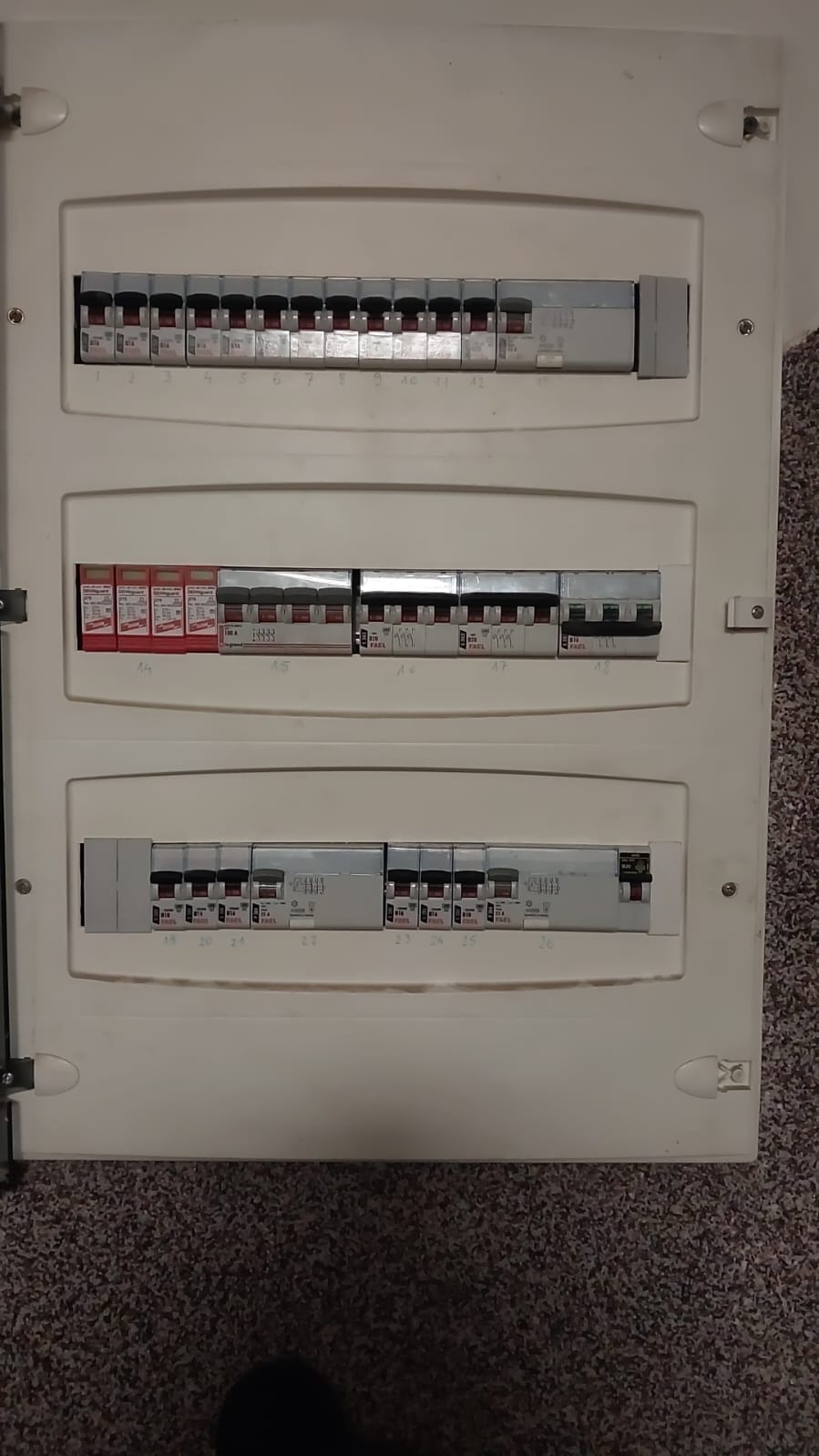 Otwarta skrzynka bezpiecznikowa z trzema rzędami wyłączników, widoczne bezpieczniki różnicowoprądowe i nadprądowe, etykiety z numerami obwodów, tło z wykładziną dywanową.