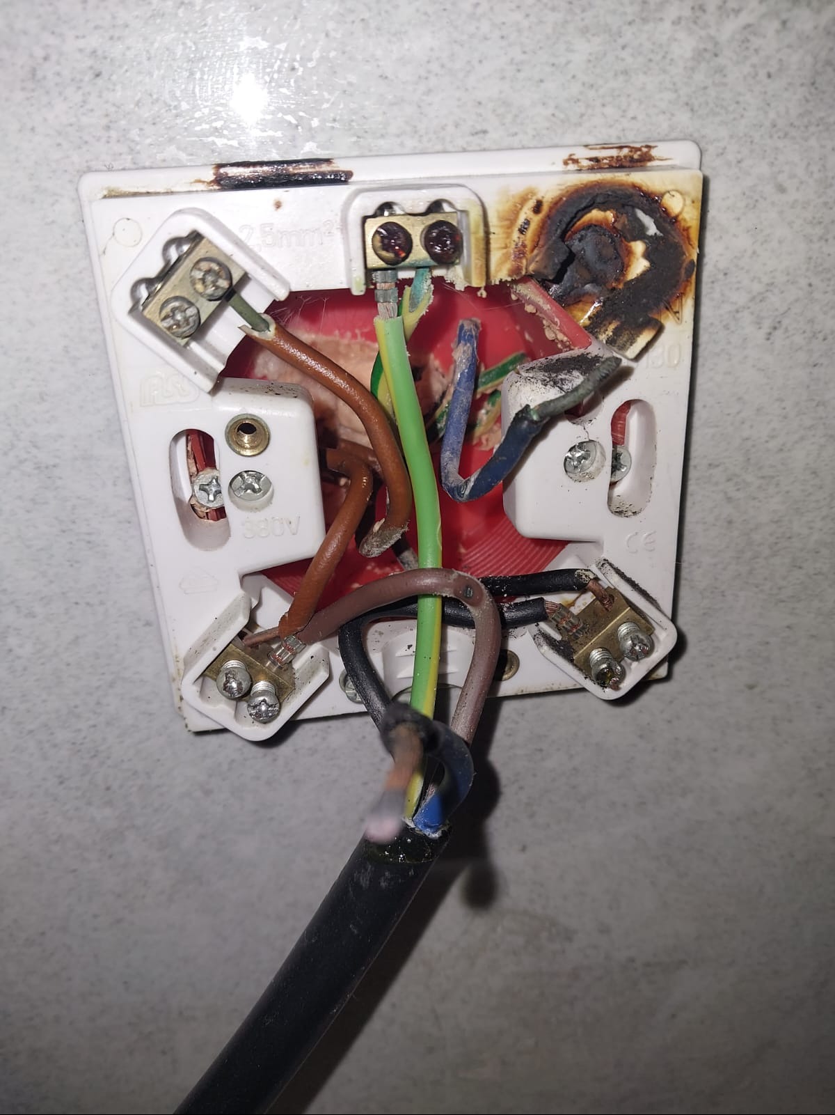 Przepalona, zwęglona instalacja elektryczna gniazdka z widocznymi przewodami i stopionym plastikiem obudowy, ślady osmalenia na ścianie.