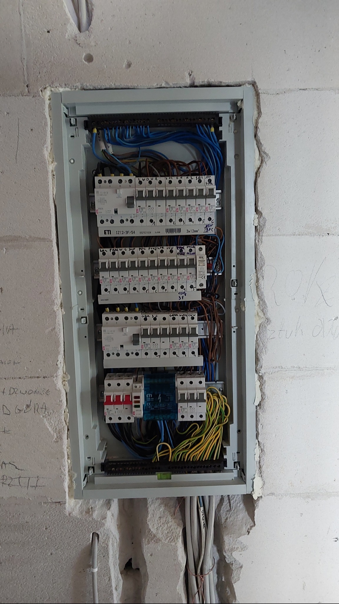 Rozdzielnica elektryczna z zainstalowanymi bezpiecznikami i okablowaniem, zamontowana w surowej ścianie z widocznymi śladami po instalacji, w trakcie prac wykończeniowych.