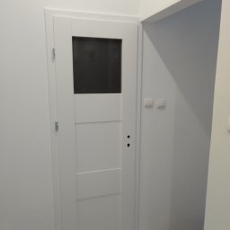 W.K Remonty i Wykończenia Wnętrz - Białe, częściowo przeszklone drzwi wewnętrzne z widocznymi zawiasami i otworami na klamkę, osadzone w świeżo pomalowanej na biało ścianie, obok fragmentu otwartego przejścia do innego pomieszczenia...