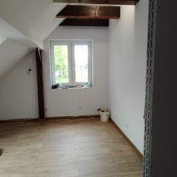 W.K Remonty i Wykończenia Wnętrz - Poddasze po remoncie z drewnianymi elementami konstrukcyjnymi, jasnymi ścianami i panelami podłogowymi, widok na okno z widokiem na zieleń, w rogu wiaderko z narzędziami malarskimi.