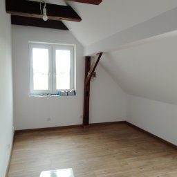 W.K Remonty i Wykończenia Wnętrz - Poddasze po remoncie: świeżo pomalowane białe ściany, ułożone panele podłogowe, widoczne drewniane belki stropowe i słup, okno z parapetem