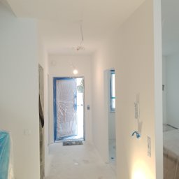 W.K Remonty i Wykończenia Wnętrz - Korytarz w trakcie remontu: ściany świeżo pomalowane na biało, zabezpieczone folią drzwi wejściowe, widoczne kable oświetleniowe pod sufitem i lampa podłogowa.