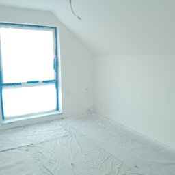 W.K Remonty i Wykończenia Wnętrz - Pokój na poddaszu po malowaniu na biało, okno zabezpieczone niebieską taśmą malarską, podłoga zakryta folią ochronną, widoczny kabel zwisający z sufitu.