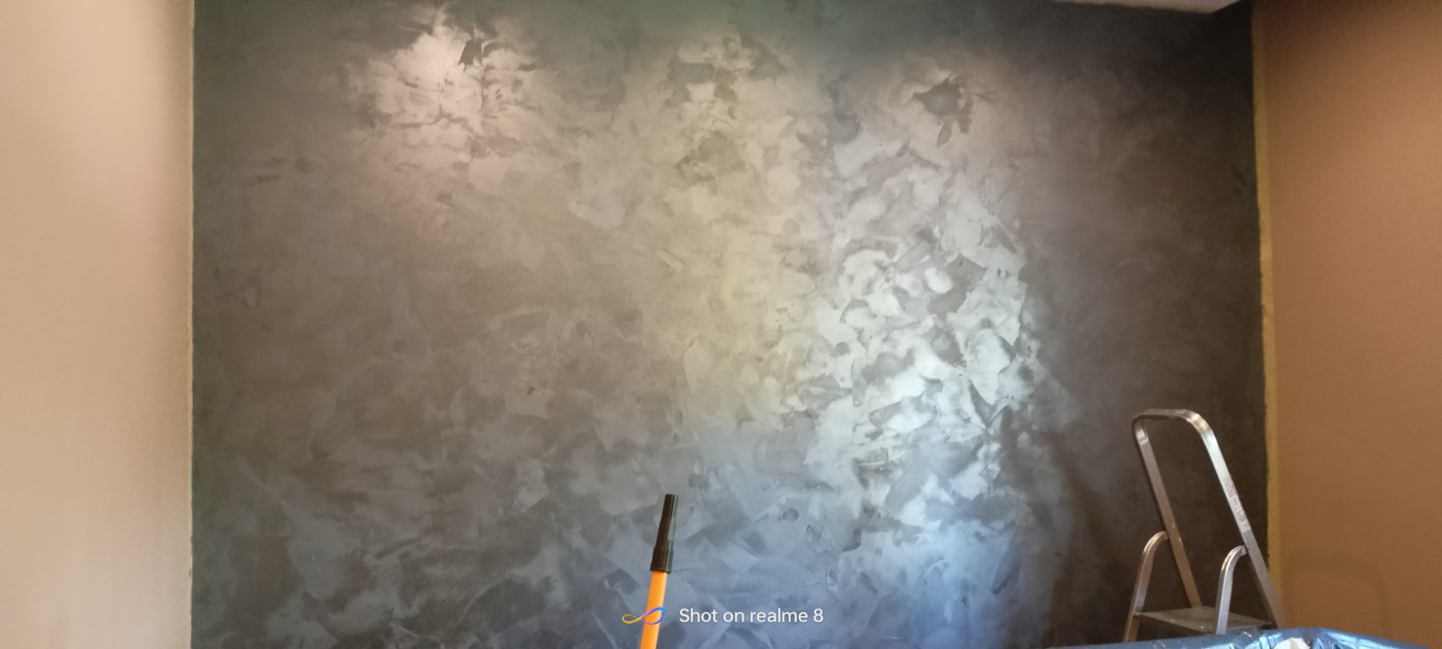 Ściana z efektem dekoracyjnym w odcieniach szarości w Szczecinie, widoczna drabina malarska i fragment ściany w kolorze beżowym.