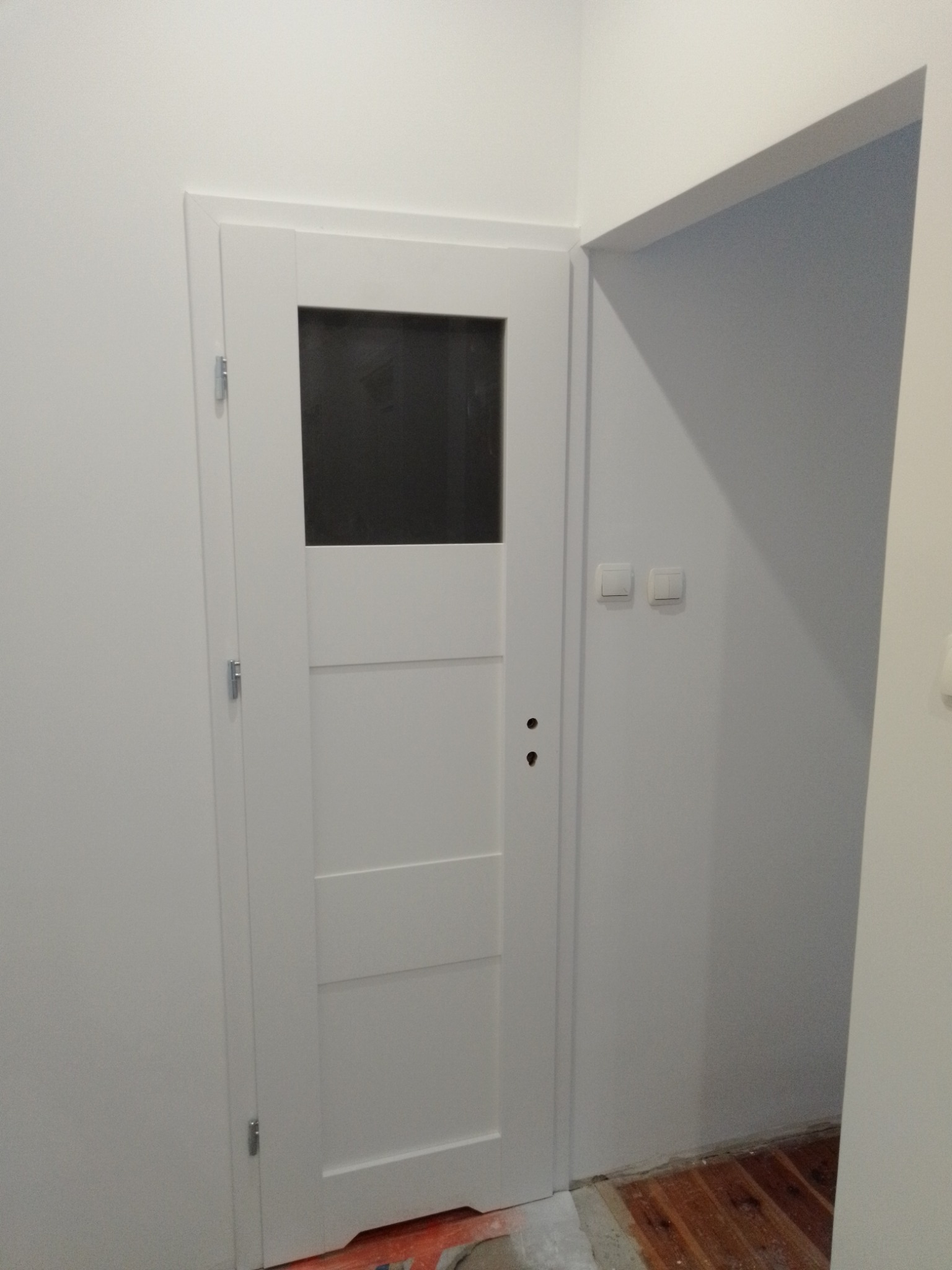 Białe, częściowo przeszklone drzwi wewnętrzne z widocznymi zawiasami i otworami na klamkę, osadzone w świeżo pomalowanej na biało ścianie, obok fragmentu otwartego przejścia do innego pomieszczenia...