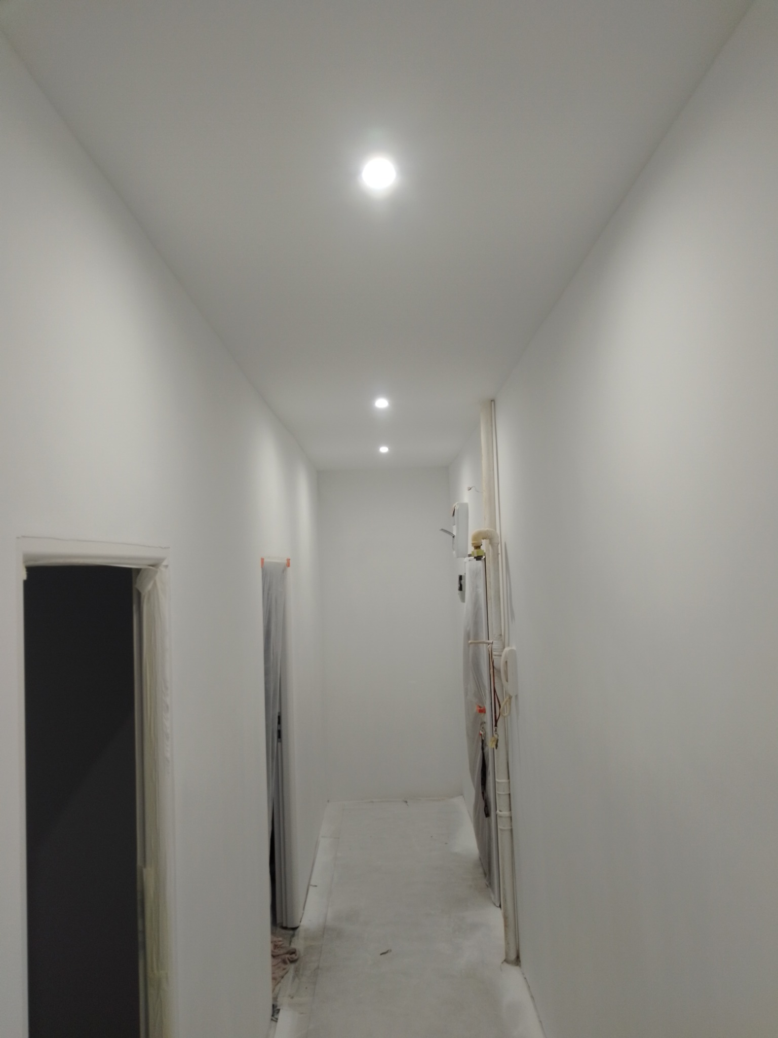 Korytarz w Szczecinie po remoncie, świeżo pomalowane na biało ściany i sufit z trzema punktowymi lampami LED, widoczne drzwi z futrynami przygotowane do montażu.