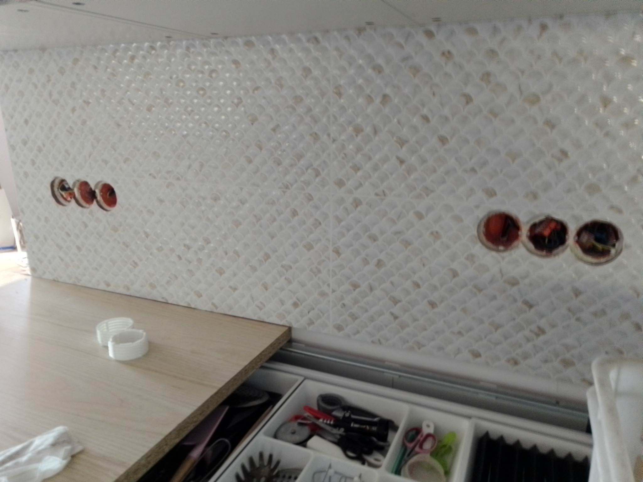 Ściana w kuchni wyłożona białymi, drobnymi płytkami o wzorze rybiej łuski, z widocznymi otworami na gniazdka elektryczne, blat roboczy z akcesoriami i otwartą szufladą z przyborami kuchennymi.