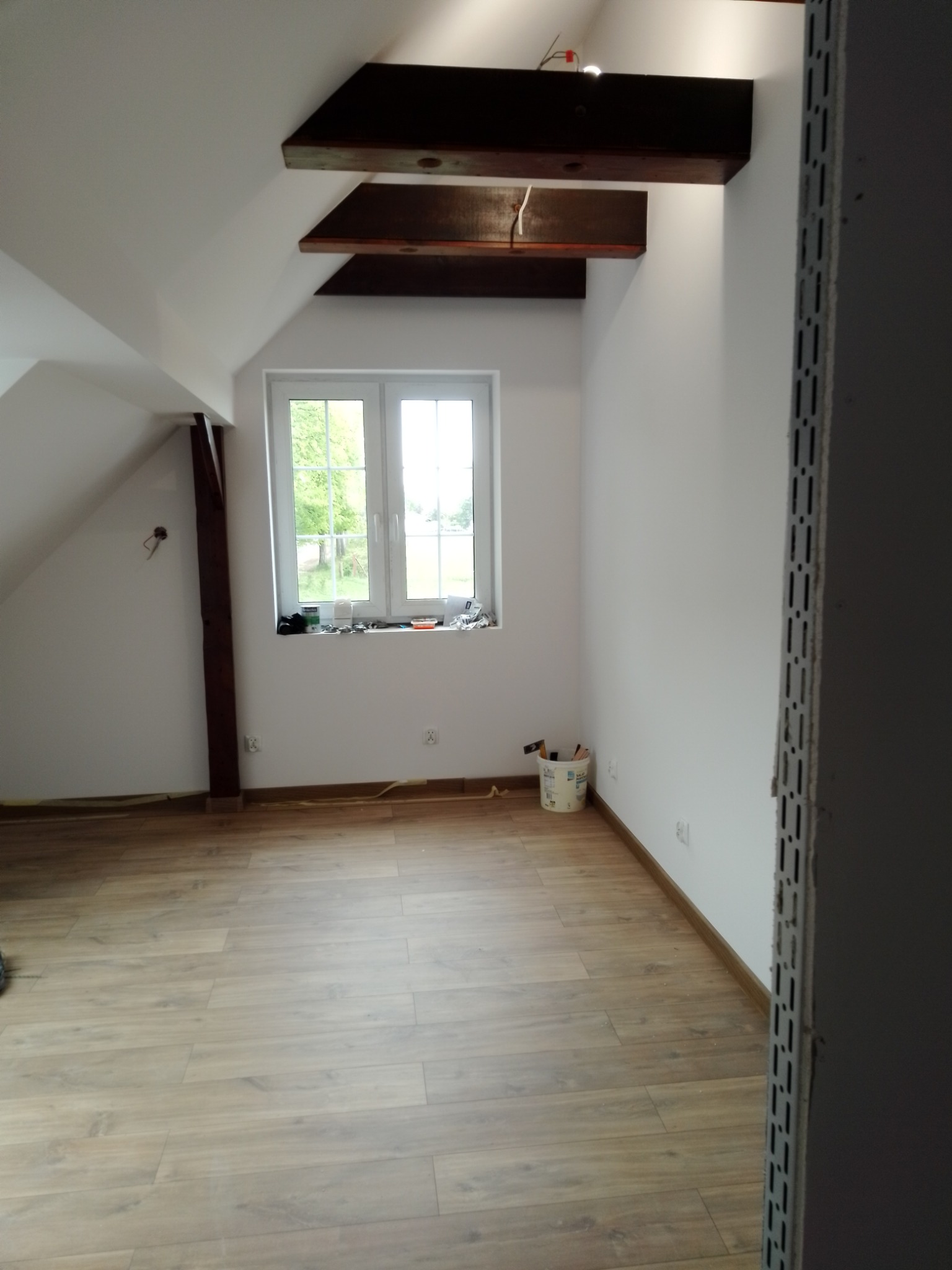 Poddasze po remoncie z drewnianymi elementami konstrukcyjnymi, jasnymi ścianami i panelami podłogowymi, widok na okno z widokiem na zieleń, w rogu wiaderko z narzędziami malarskimi.