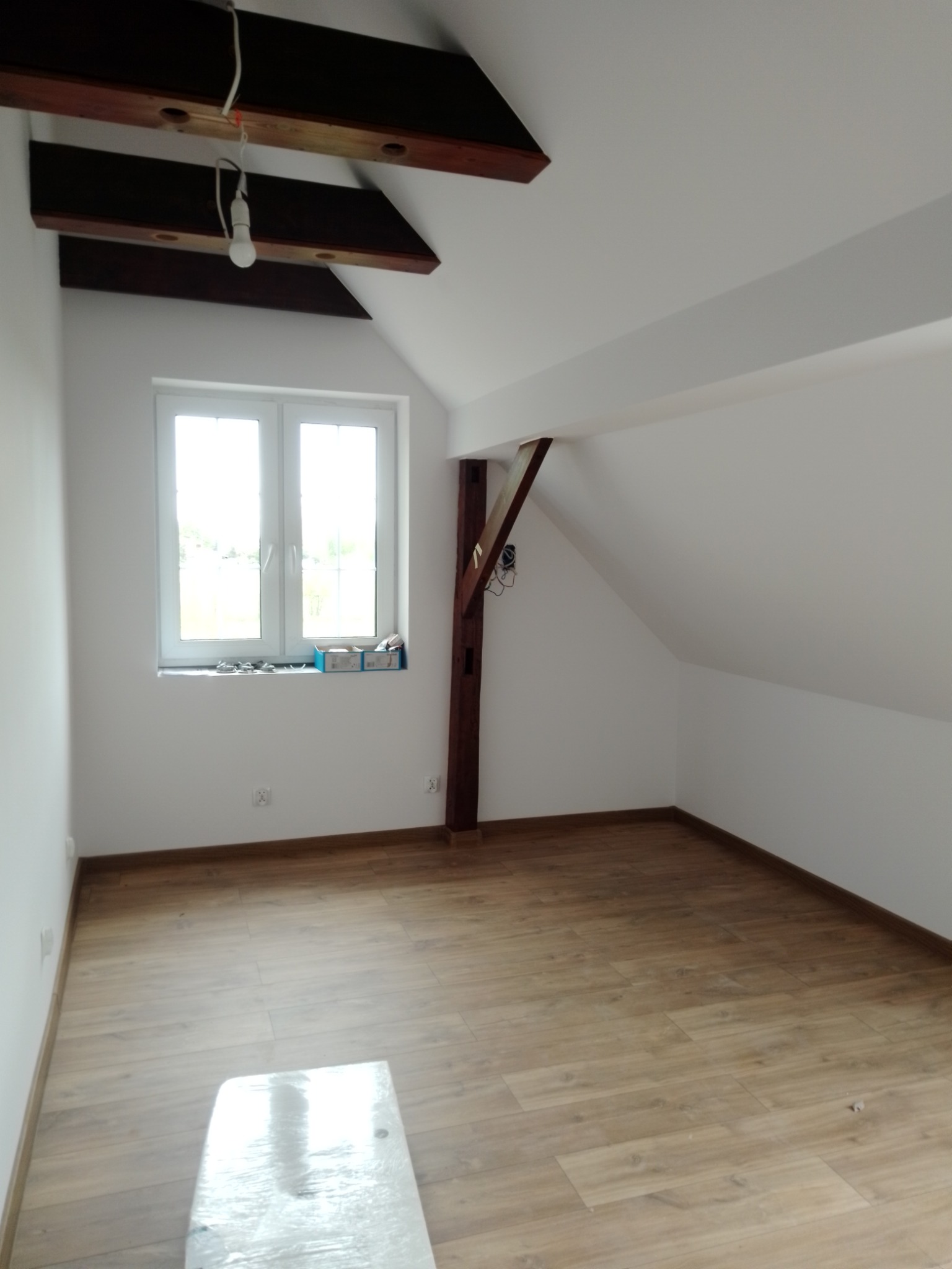 Poddasze po remoncie: świeżo pomalowane białe ściany, ułożone panele podłogowe, widoczne drewniane belki stropowe i słup, okno z parapetem