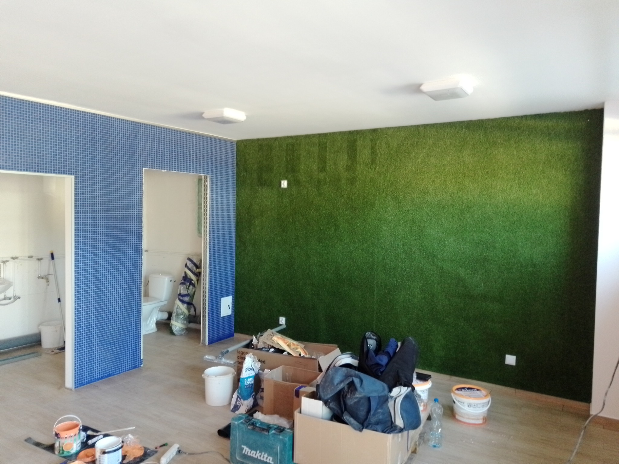 Wnętrze w trakcie remontu: ściana pokryta zieloną imitacją trawy, fragment ściany z niebieską mozaiką, widoczne narzędzia i materiały budowlane, toaleta w tle.