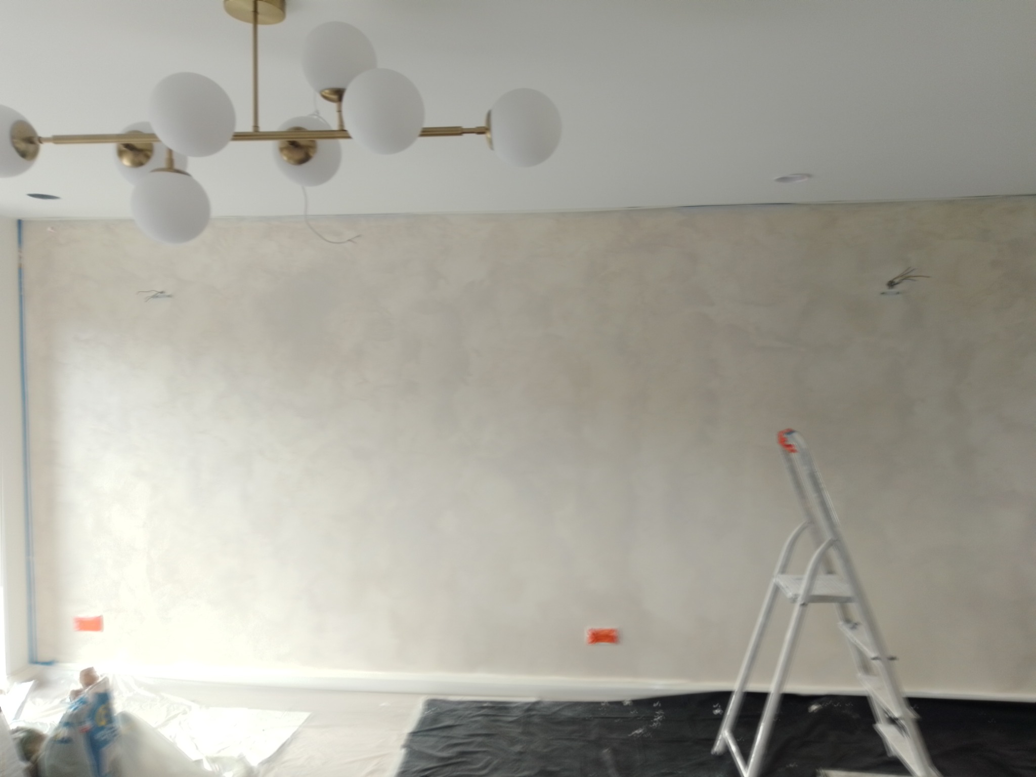 Ściana po nałożeniu gładzi w odcieniach beżu, przykryta folią ochronną, widoczna drabina aluminiowa i nowoczesna lampa sufitowa z kulistymi kloszami.