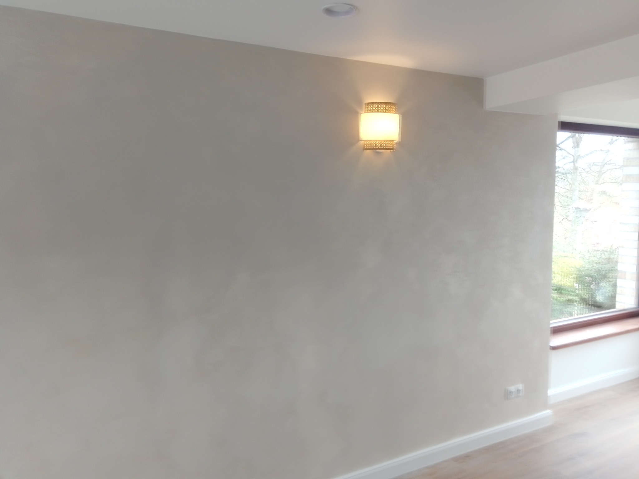 Ściana w pokoju pokryta gładzią w odcieniach szarości, oświetlona kinkietem z ażurowym abażurem, widoczne okno z drewnianym parapetem i fragment podłogi.
