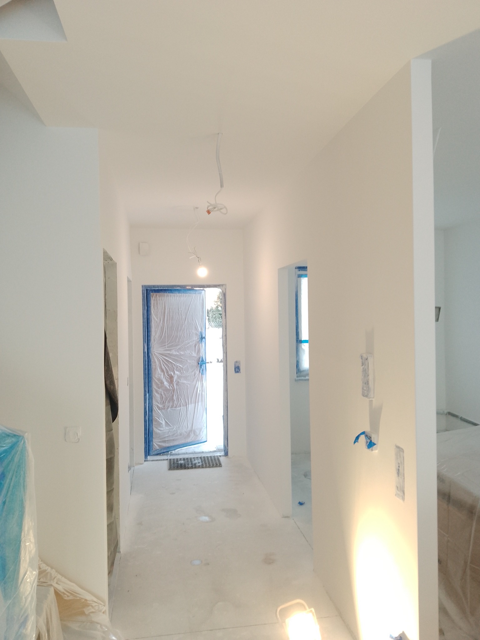 Korytarz w trakcie remontu: ściany świeżo pomalowane na biało, zabezpieczone folią drzwi wejściowe, widoczne kable oświetleniowe pod sufitem i lampa podłogowa.