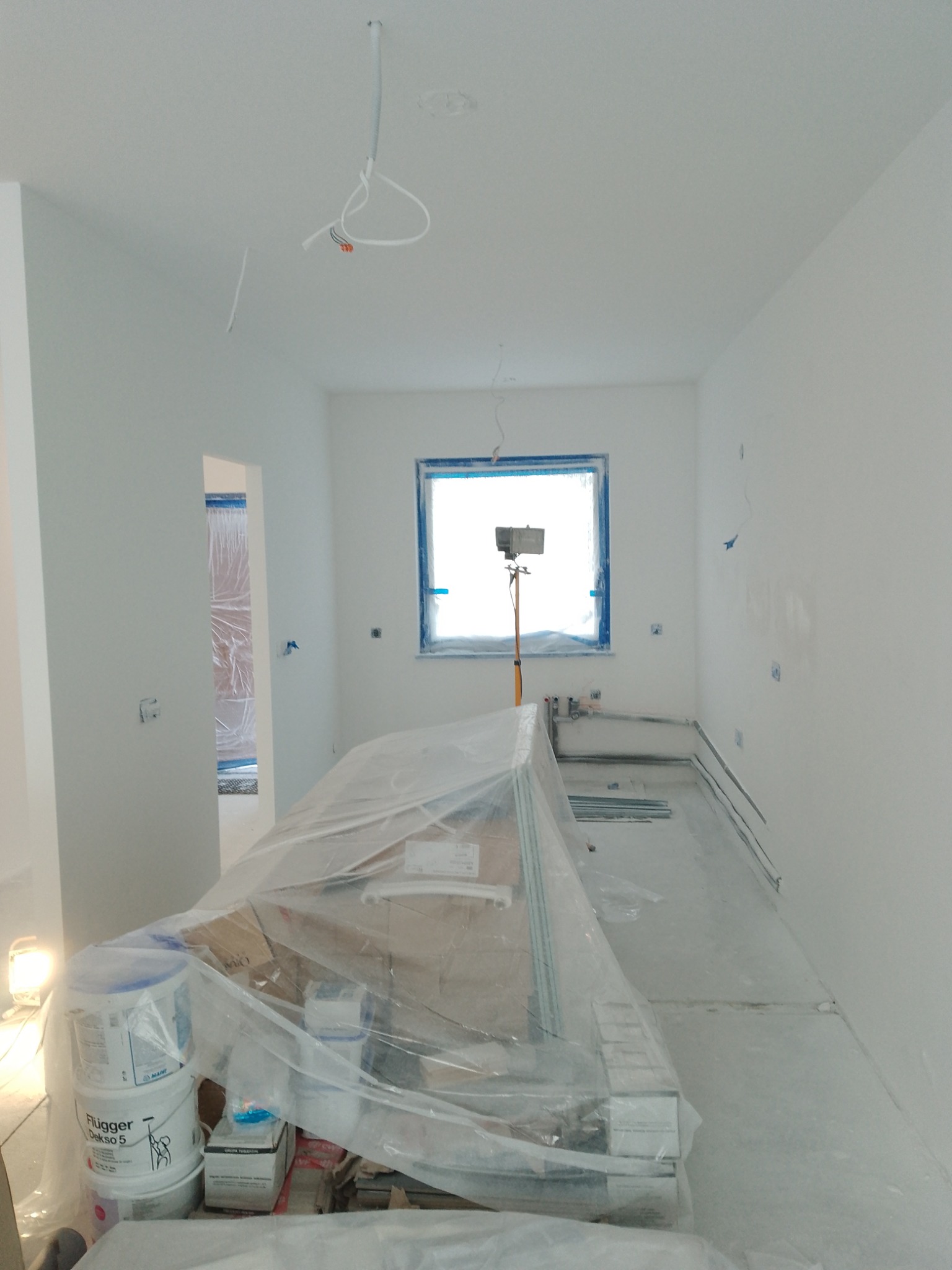 Wnętrze w trakcie prac wykończeniowych: świeżo pomalowane białe ściany, okno zabezpieczone taśmą malarską, widoczne puszki z farbą Flügger Dekso 5 i narzędzia pod folią ochronną, przewody...