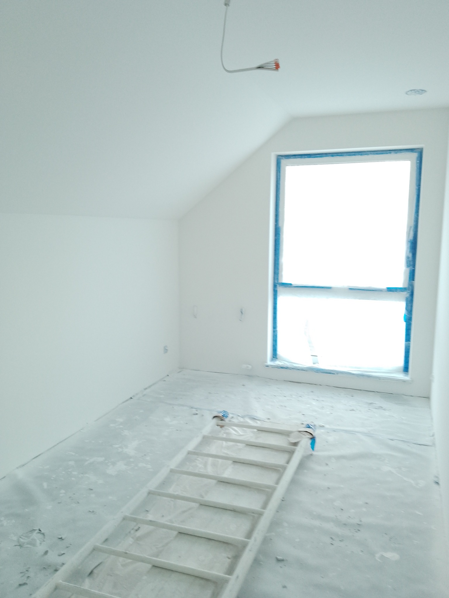 Poddasze po świeżym pomalowaniu na biało, zabezpieczone okno taśmą malarską, rozłożona drabina na folii ochronnej, przygotowane do dalszych prac wykończeniowych.