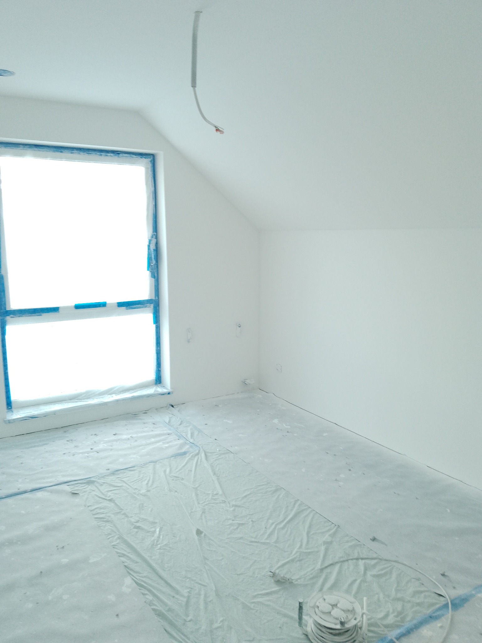 Pokój na poddaszu po malowaniu na biało, okno zabezpieczone niebieską taśmą malarską, podłoga zakryta folią ochronną, widoczny kabel zwisający z sufitu.