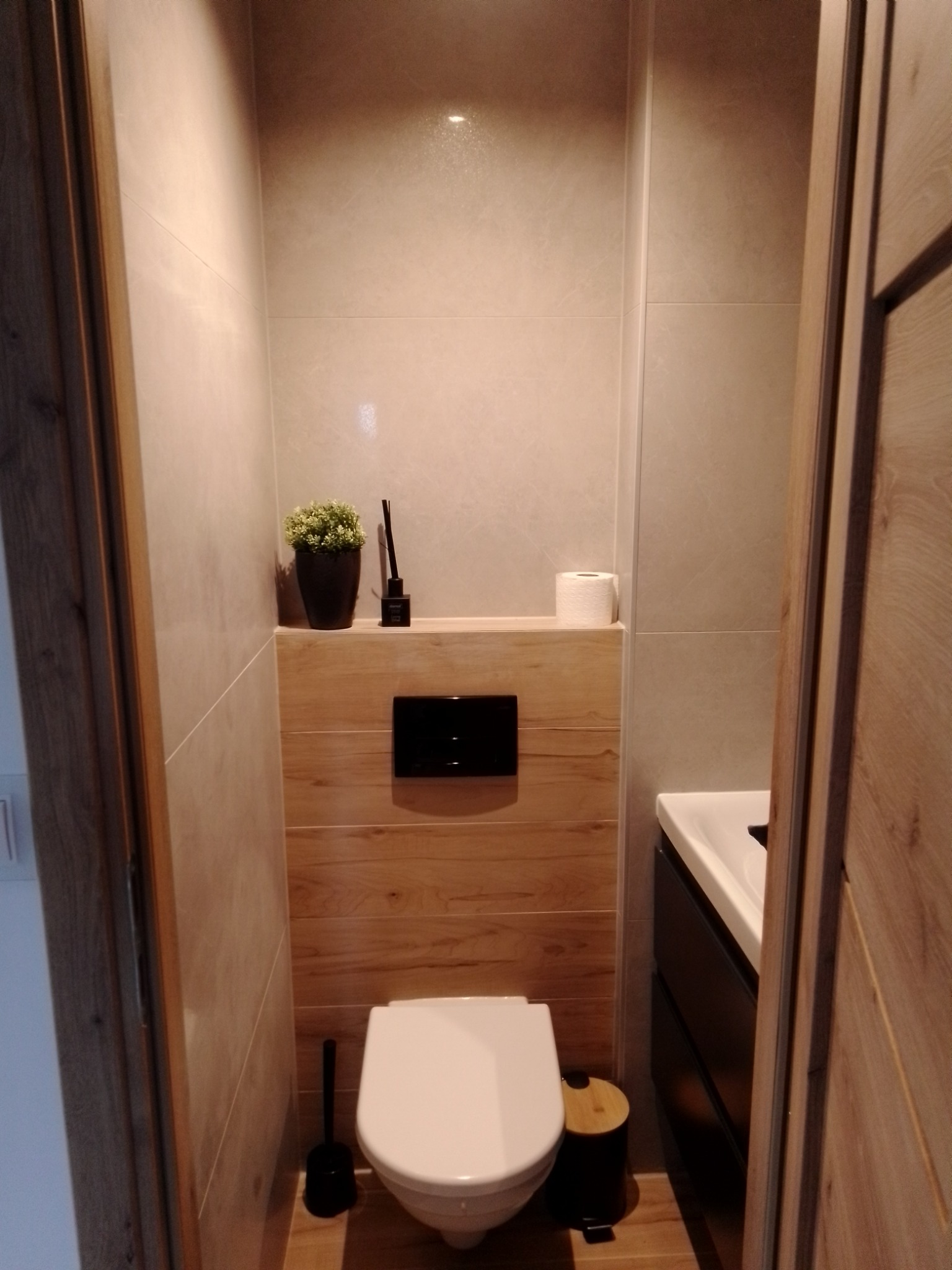 Nowoczesna toaleta z podwieszaną miską WC, drewnianą zabudową stelaża, szarą ceramiką ścienną oraz umywalką z czarną szafką, widziana z perspektywy wejścia przez drewniane drzwi.