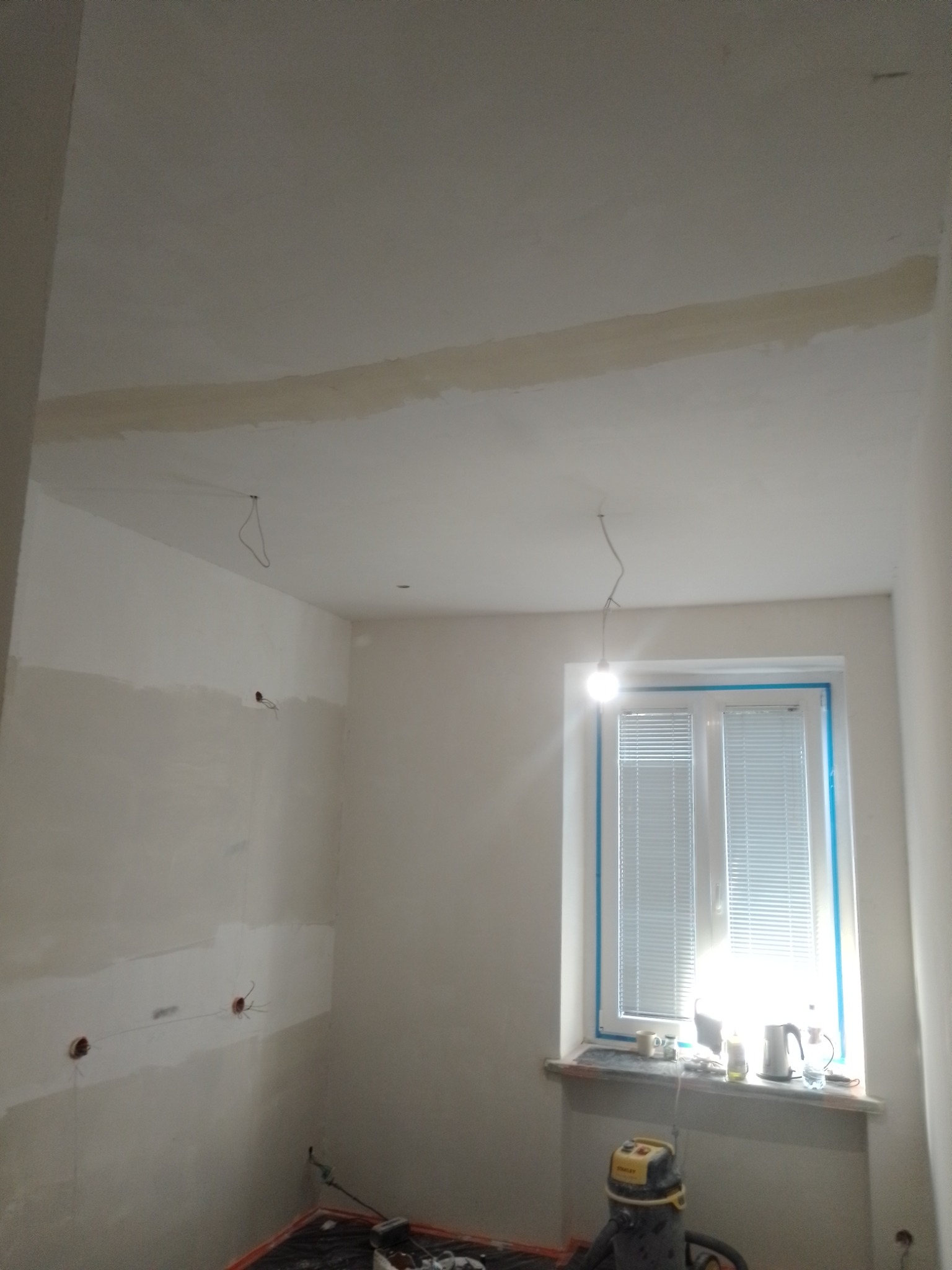 Wnętrze pokoju w trakcie remontu: ściany częściowo pokryte gładzią, okno zabezpieczone taśmą, widoczne przewody elektryczne i odkurzacz przemysłowy.