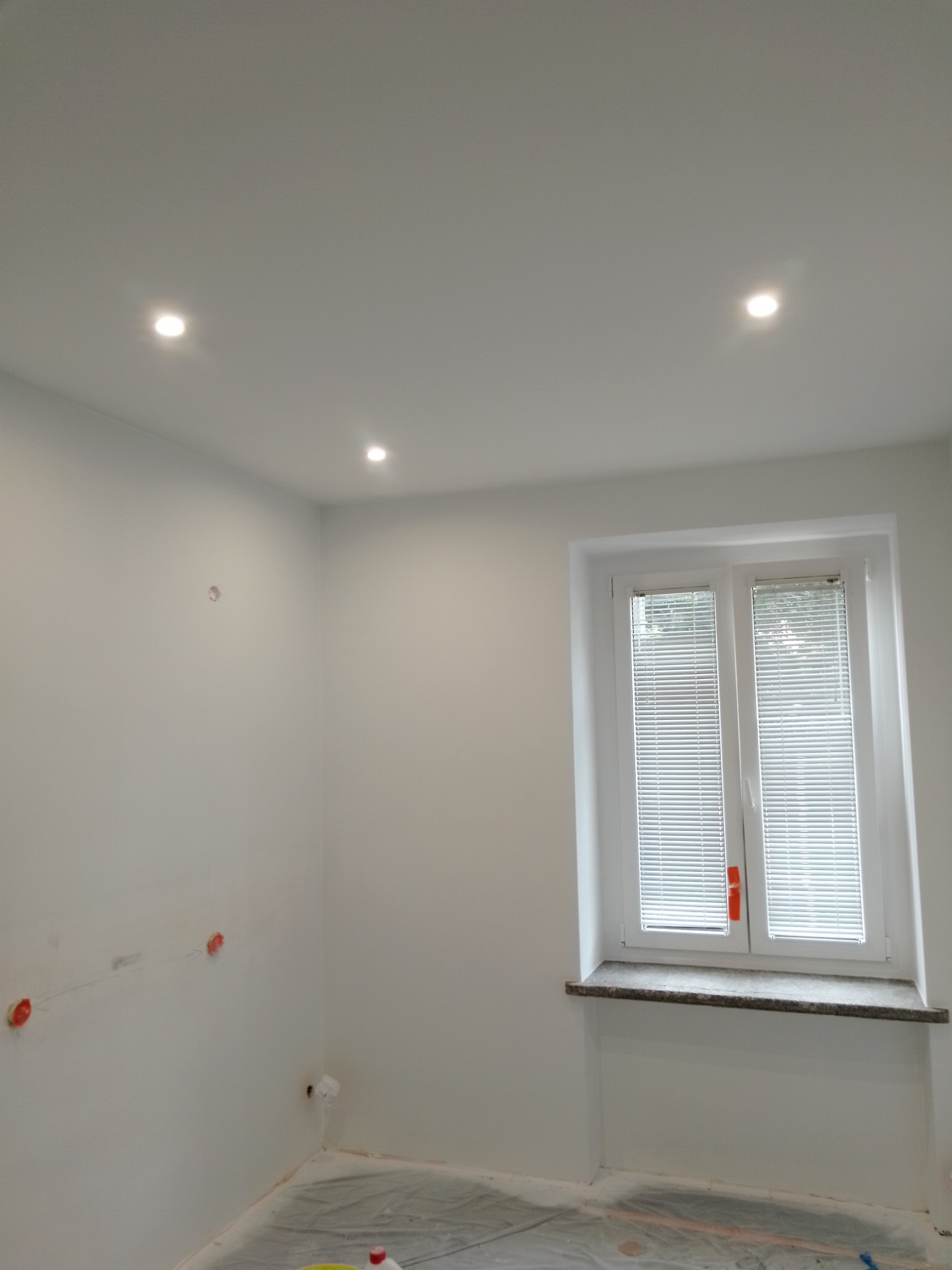 Pokój po malowaniu na biało z trzema punktowymi lampami w suficie, zabezpieczoną folią podłogą i oknem z białymi żaluzjami.