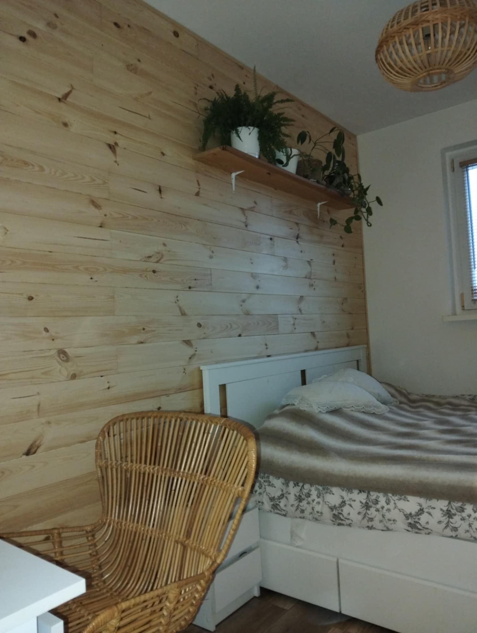 Sypialnia z drewnianą ścianą wyłożoną panelami, rattanowym fotelem i półką z roślinami, oświetlona lampą z wikliny.