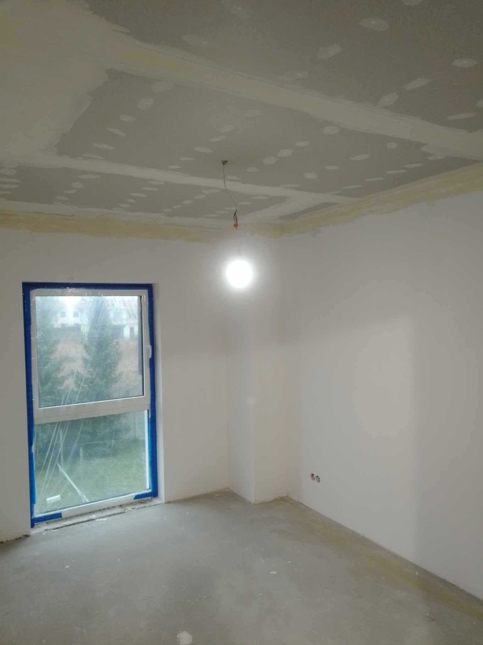 Pokój w trakcie przygotowania do malowania: surowy sufit z widocznymi łatami gładzi, białe ściany, okno zabezpieczone niebieską taśmą malarską, betonowa podłoga, wisząca żarówka.