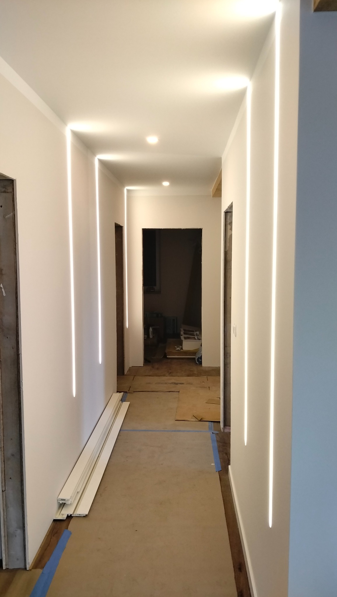 Korytarz z białymi ścianami i pionowymi, wpuszczonymi liniami świetlnymi. Na podłodze zabezpieczenia i listwy przypodłogowe. Widoczne otwarte przejścia do innych pomieszczeń.