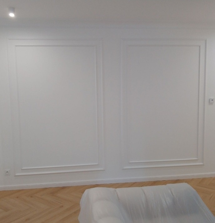 Białe ściany z dekoracyjnymi listwami w prostokątnych ramach, parkiet w jodełkę, częściowo zakryty mebel folią ochronną. Minimalistyczne wykończenie wnętrza.