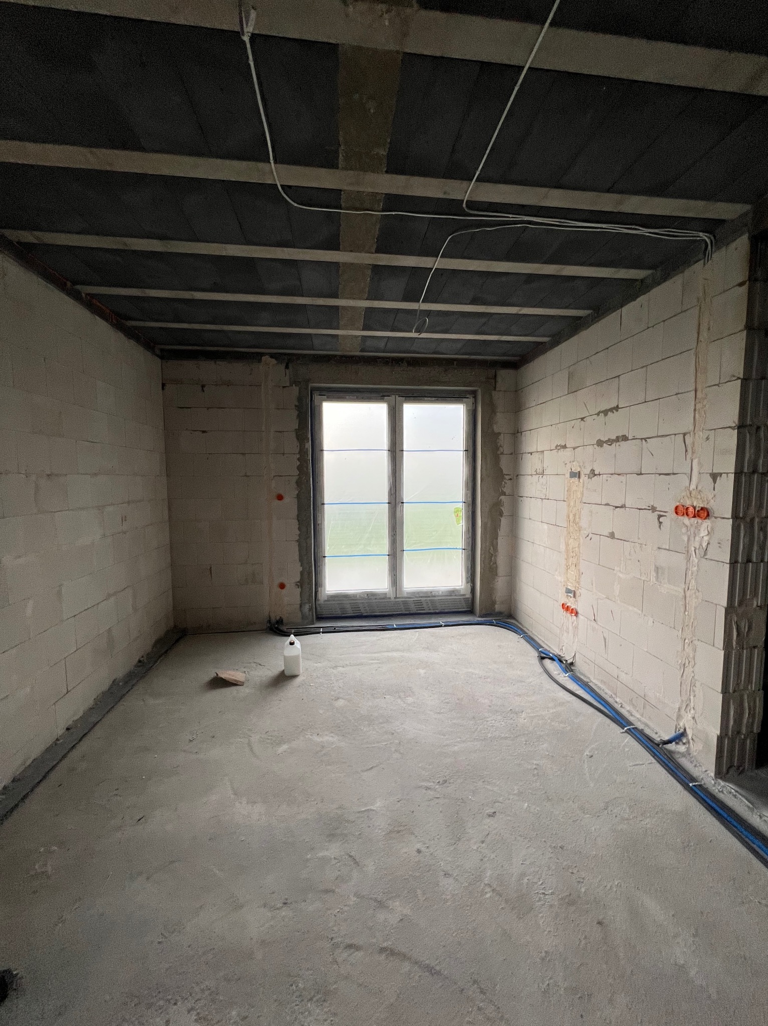 Surowy stan wnętrza: ściany z betonu komórkowego, okno z folią ochronną, instalacja elektryczna w peszlach przy podłodze i gniazda w ścianie. W tle widok na zewnątrz.